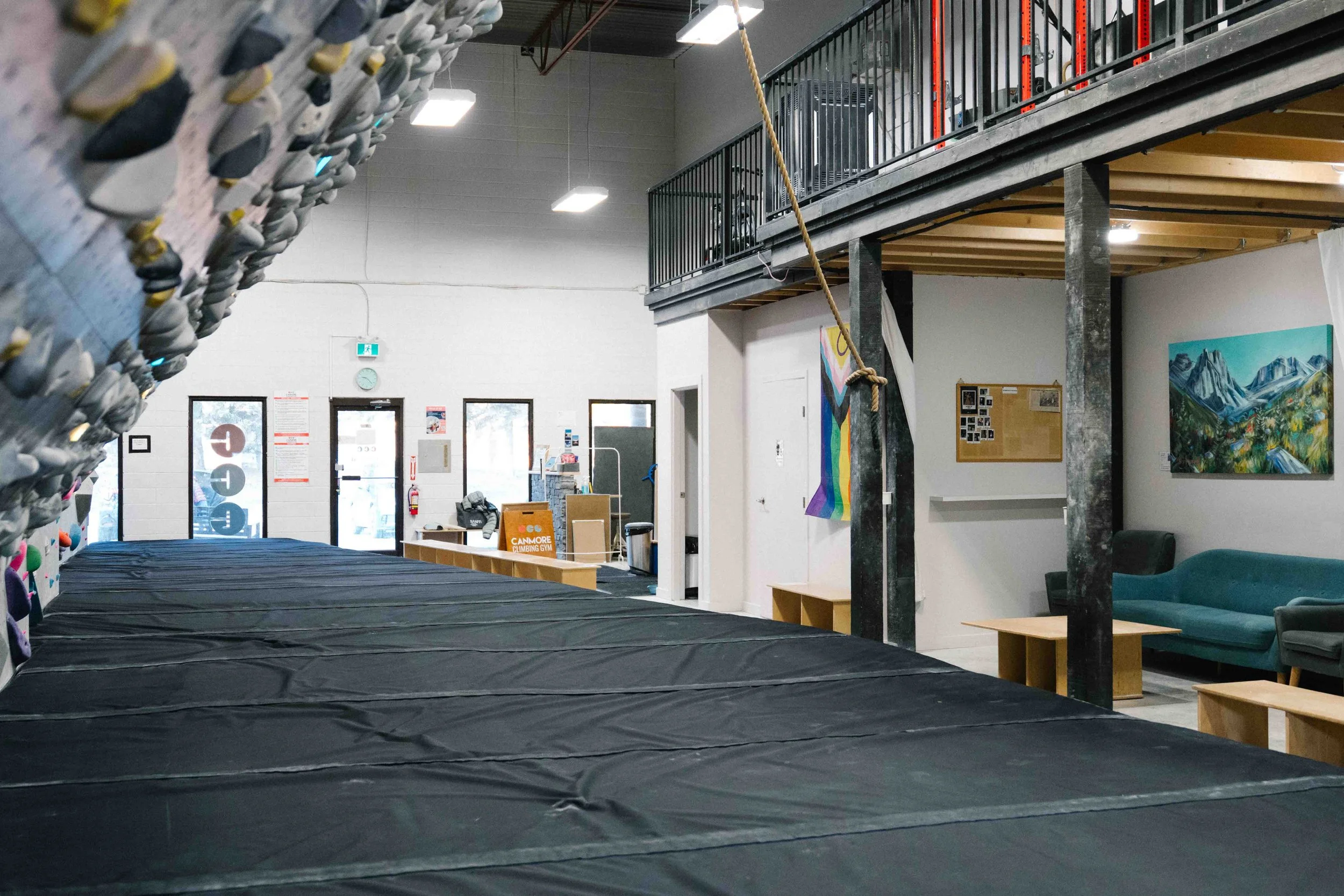 Indoor climbing gym with a large array of climbing holds on the left wall, padded flooring, a climbing area, and seating area in the background, with a lofted area overhead and artwork on the walls.