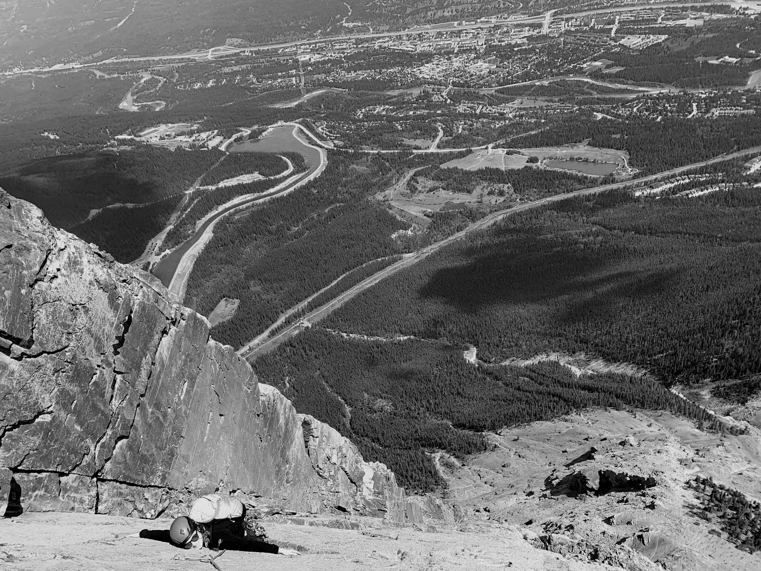 Climber resting on a rocky mountain ledge overlooking a valley with winding roads, forests, and a town in the distance. The climber is lying on the ground with a helmet beside them.