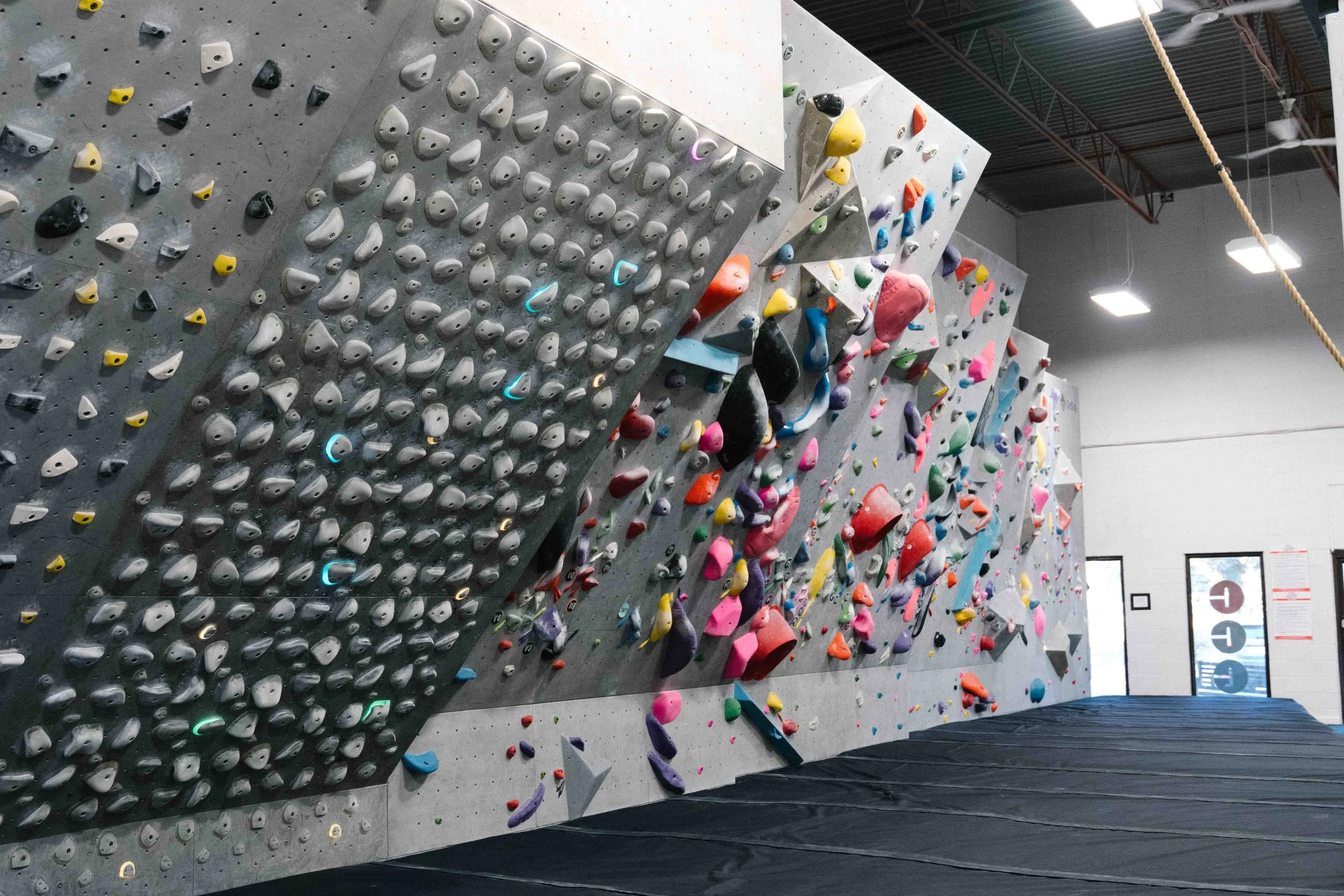 Indoor bouldering gym wall with various colorful climbing holds of different shapes and sizes, climbing pads on the floor, and ceiling lights.