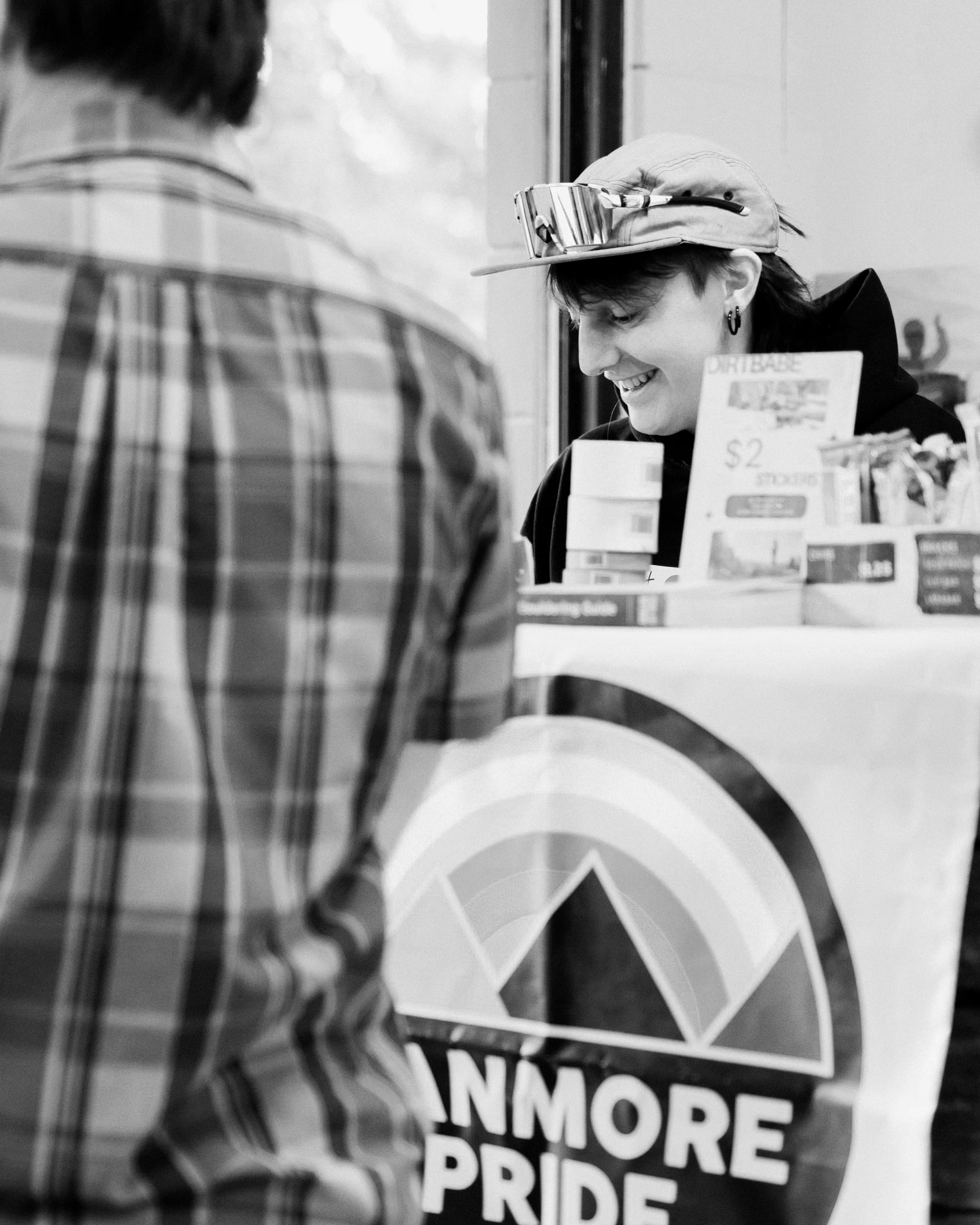 A person with dark hair wearing earrings and a cap with sunglasses on it smiles, standing behind a display of rainbow-colored arches and a sign that reads 'More Pride' in black and white.