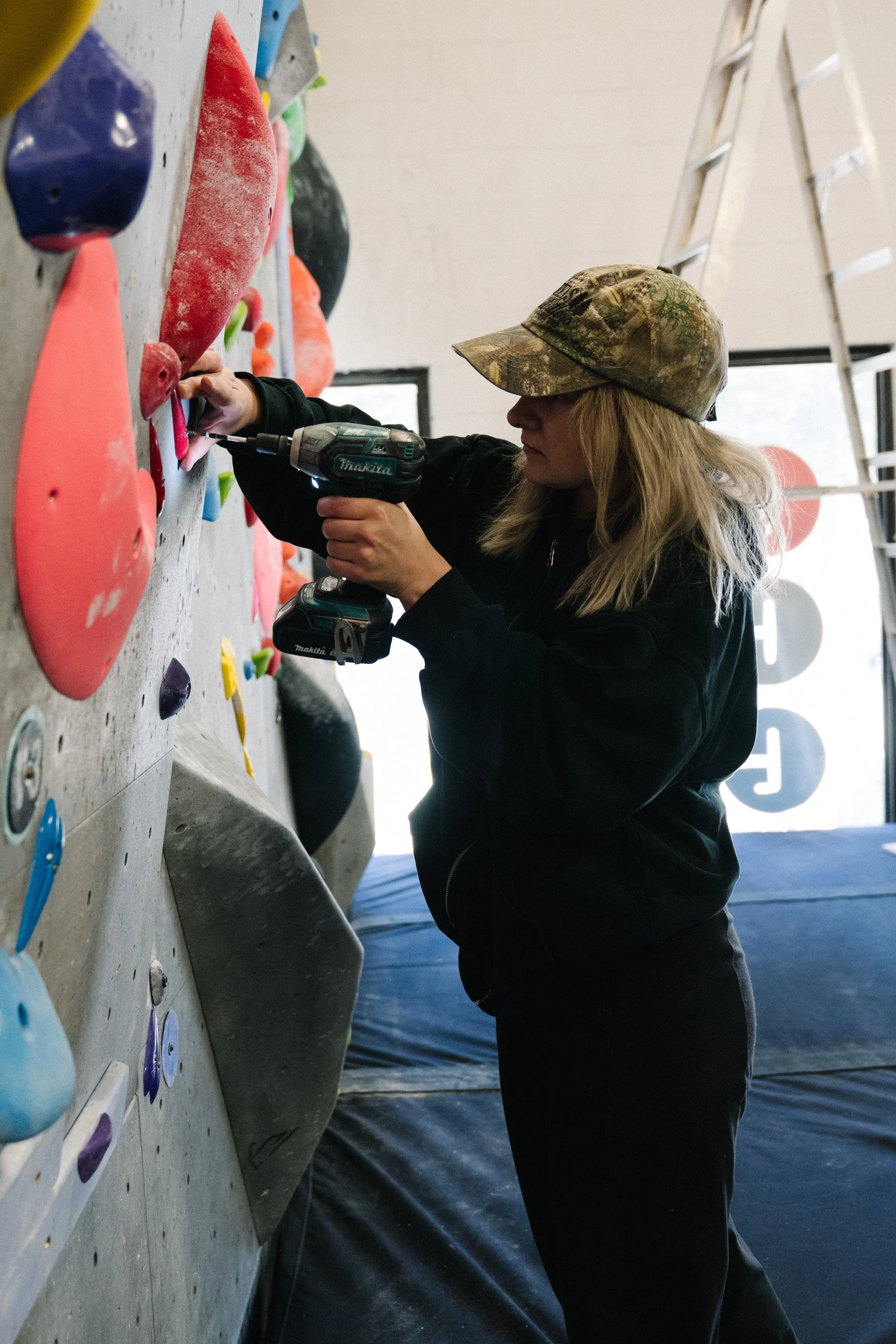 A woman with blonde hair wearing a camouflage cap and black jacket is using a power drill to tighten a bolt on an indoor rock climbing wall with various colored holds.