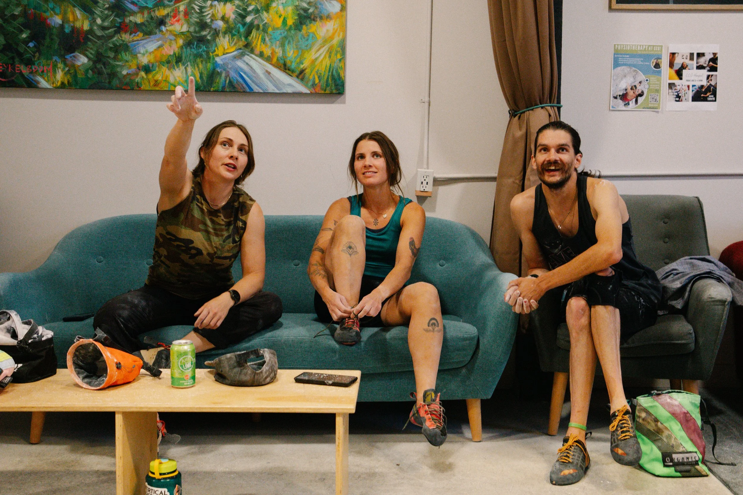 Three people sitting on a blue couch in a room with a colorful painting on the wall behind them. The person on the left is a woman with brown hair pointing upwards. The middle person is a woman with tattoos on her arms and legs, sitting with her legs