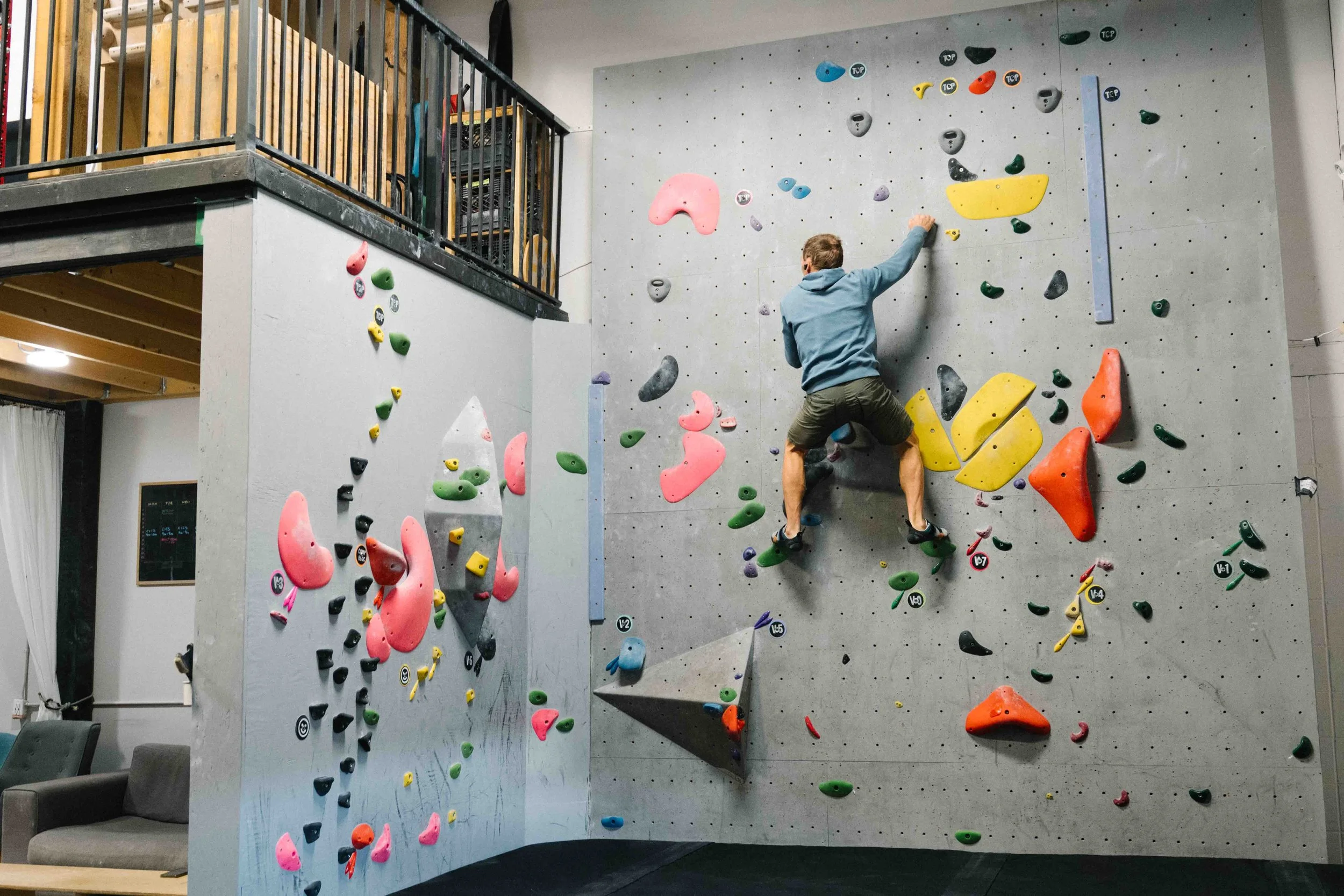 An indoor bouldering gym with colorful climbing holds on a gray wall, where a man in a blue hoodie and shorts is climbing. A seating area with chairs is visible in the lower left corner.