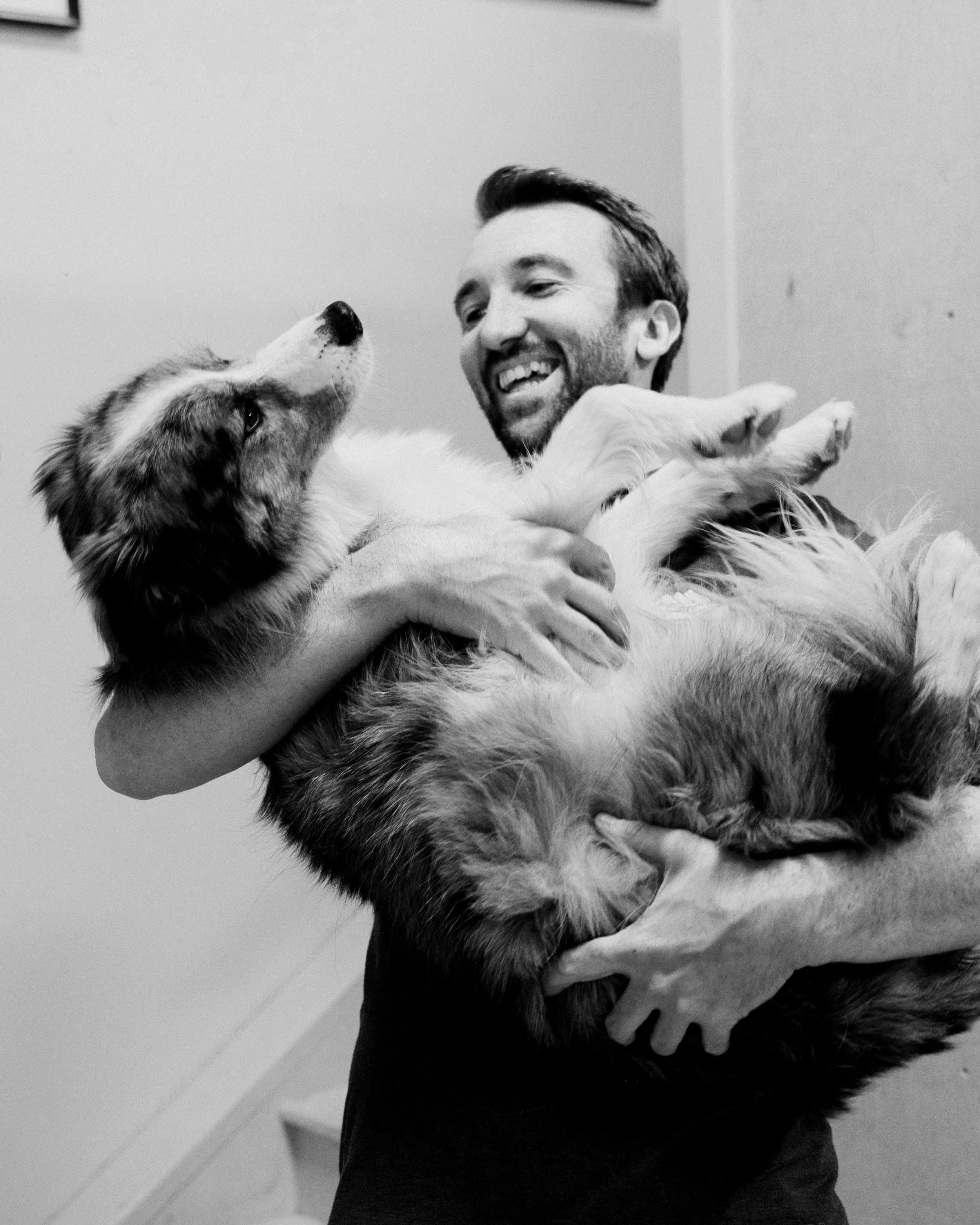A man with a beard smiling and holding a large fluffy dog with a thick coat, while the dog looks at him.