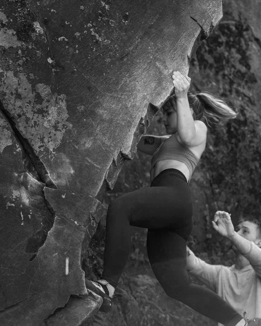 A woman rock climbing outdoors on a steep rock face, with a young girl watching her and reaching out. Black and white photo.