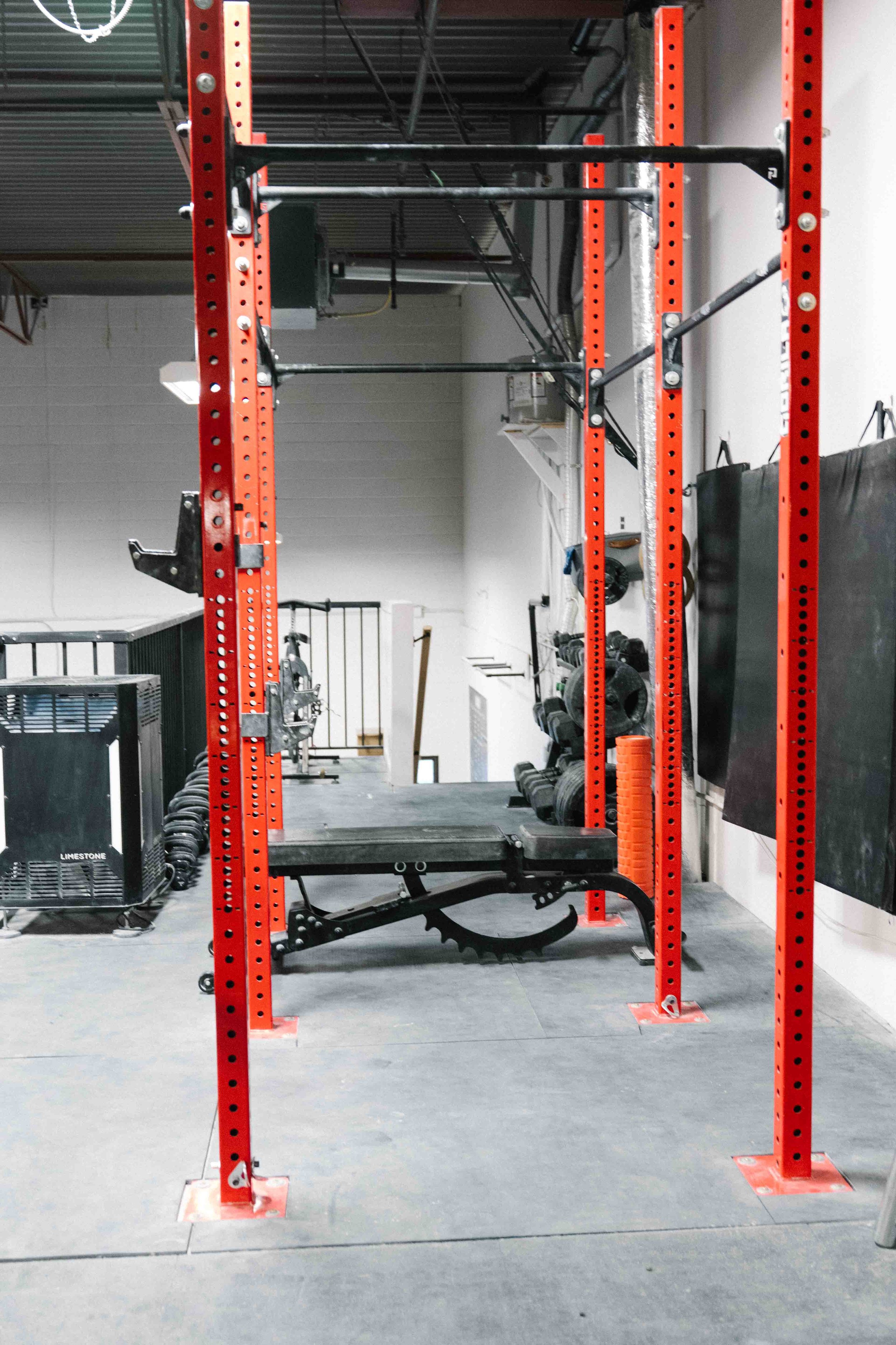 A gym with a red power rack, black weight bench, and dumbbells, with a black curtain on the right and various gym equipment in the background.