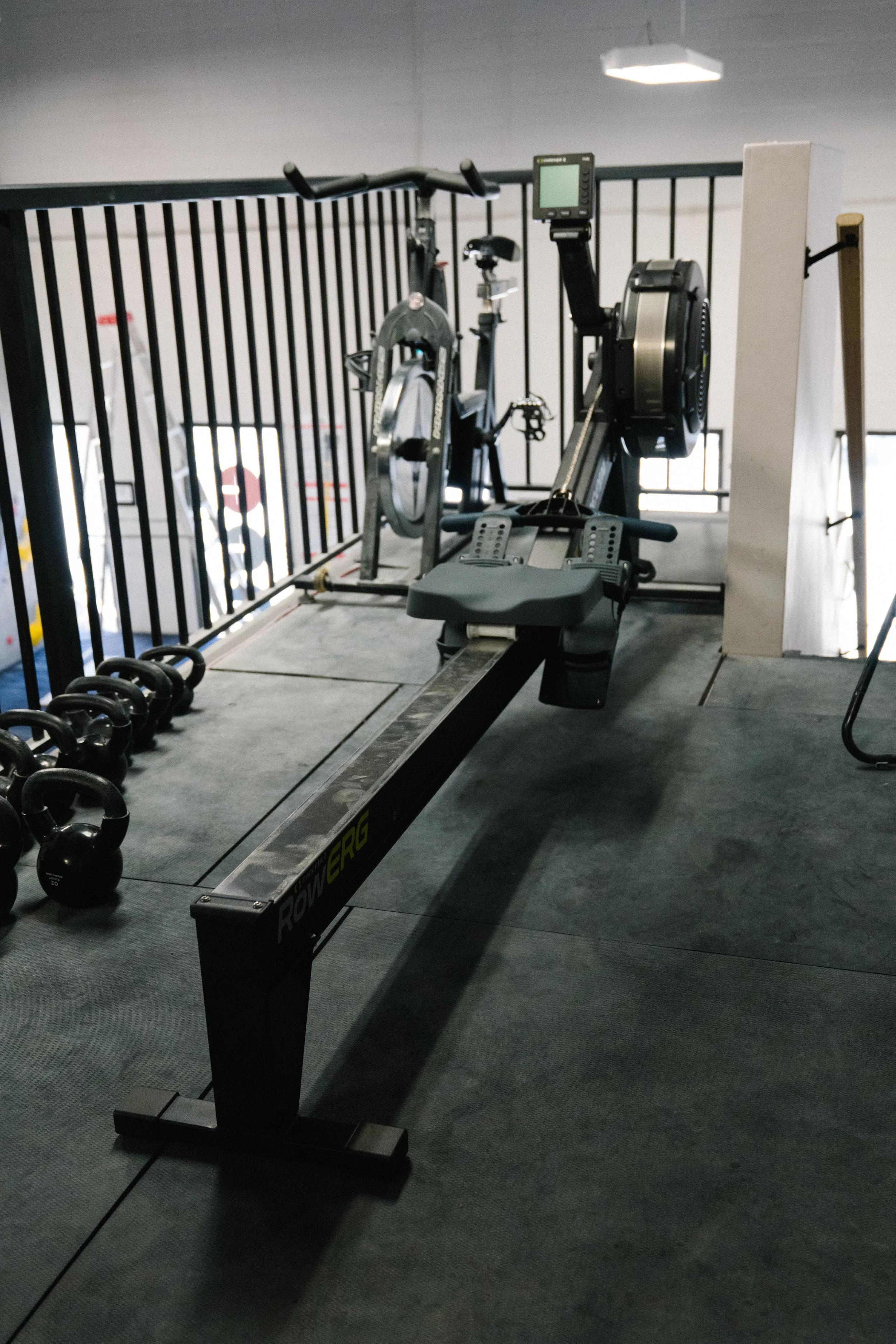 Rowing machine in a gym with kettlebells and black metal fencing in the background.