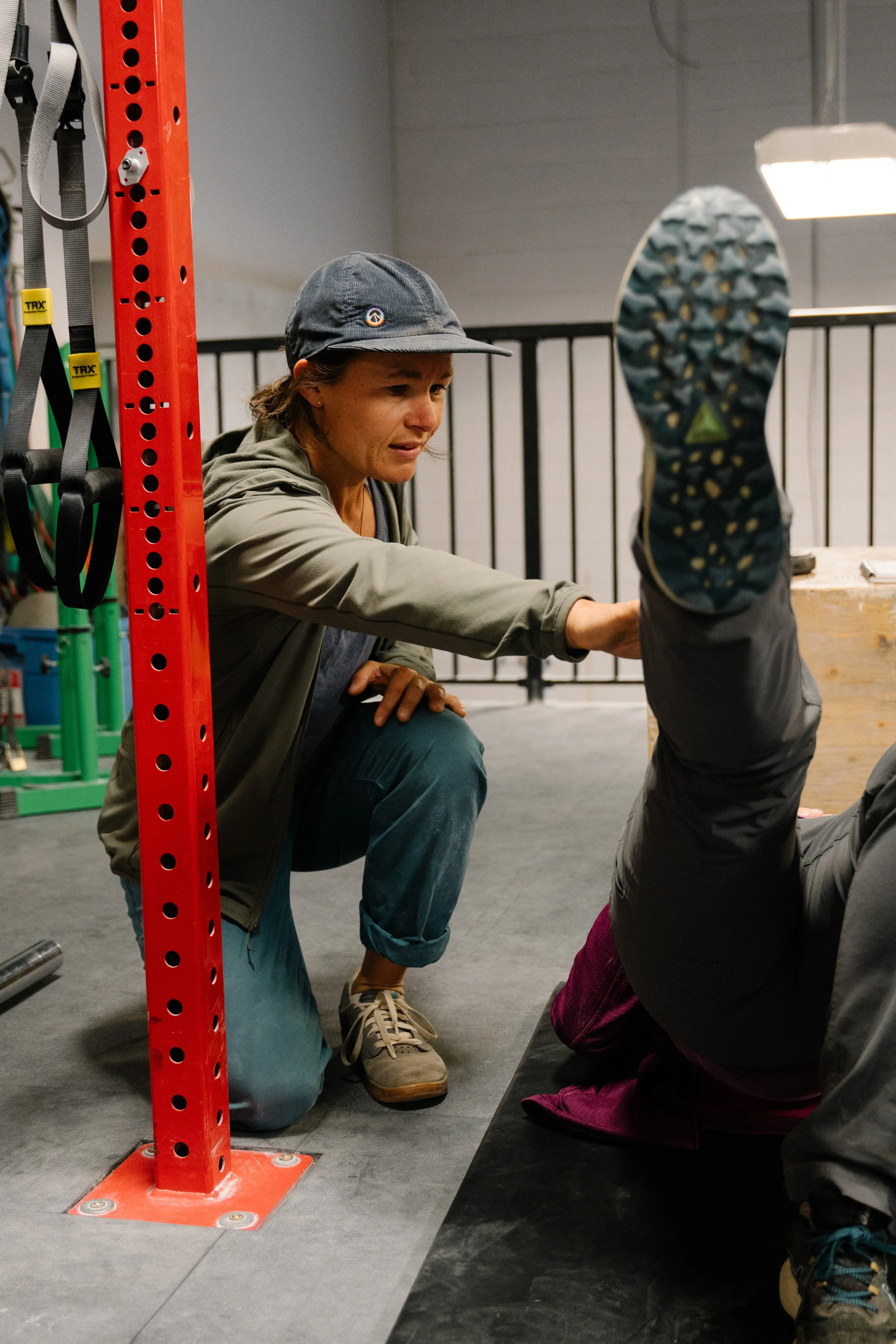 A woman assisting a person with stretching or physical therapy on a mat in a gym or fitness center, with gym equipment and a metal railing in the background.