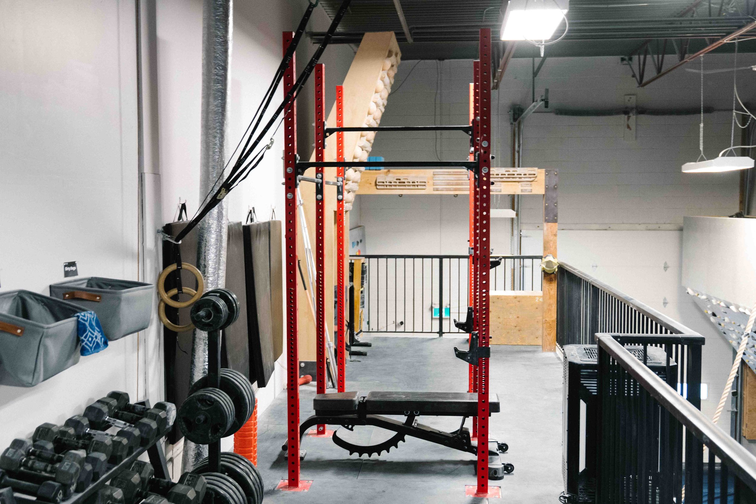 A gym or fitness workspace with equipment including dumbbells, weight plates, and a red squat rack. The space has a concrete floor, a wall on the left with storage bins and hanging accessories, and industrial ceiling with lighting and ductwork.