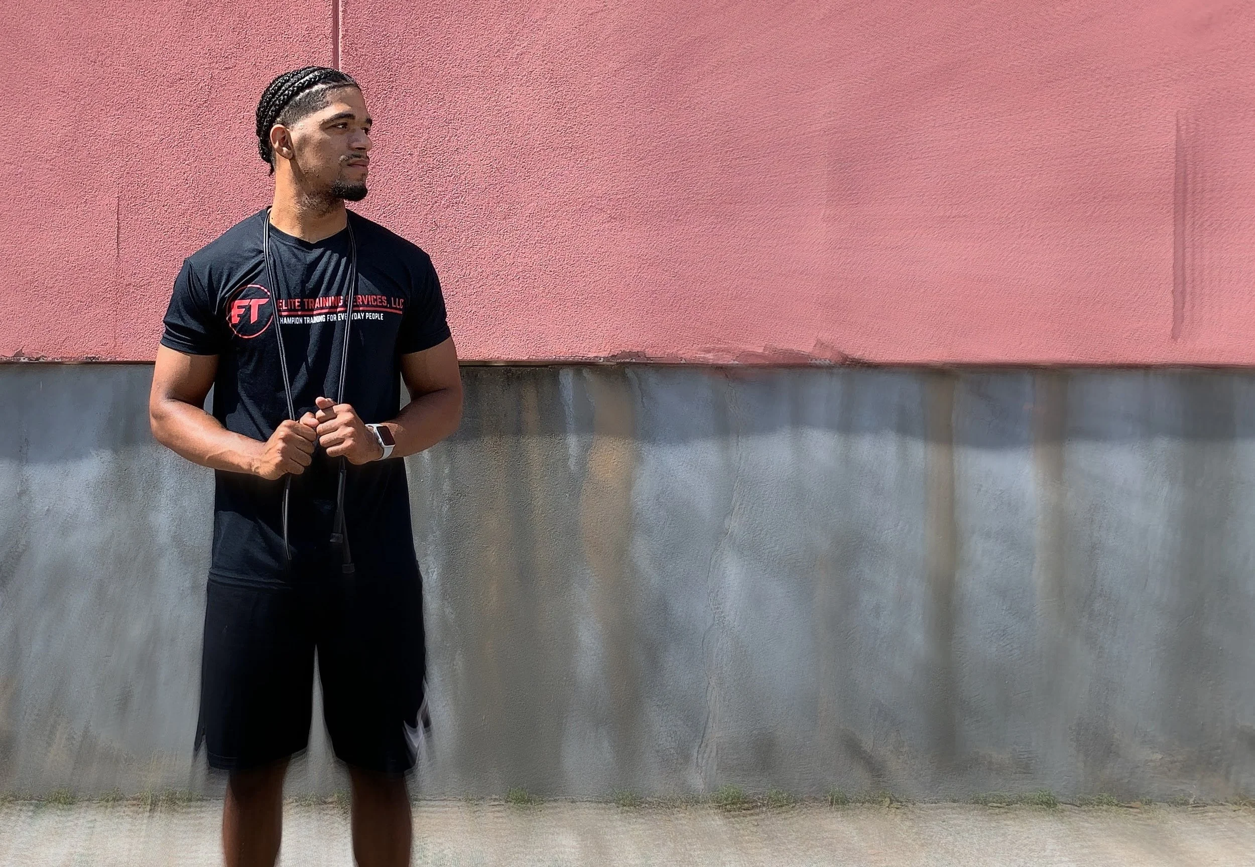 A man with braided hair wearing a black t-shirt and shorts, holding a jump rope, standing outdoors against a pink and gray wall.