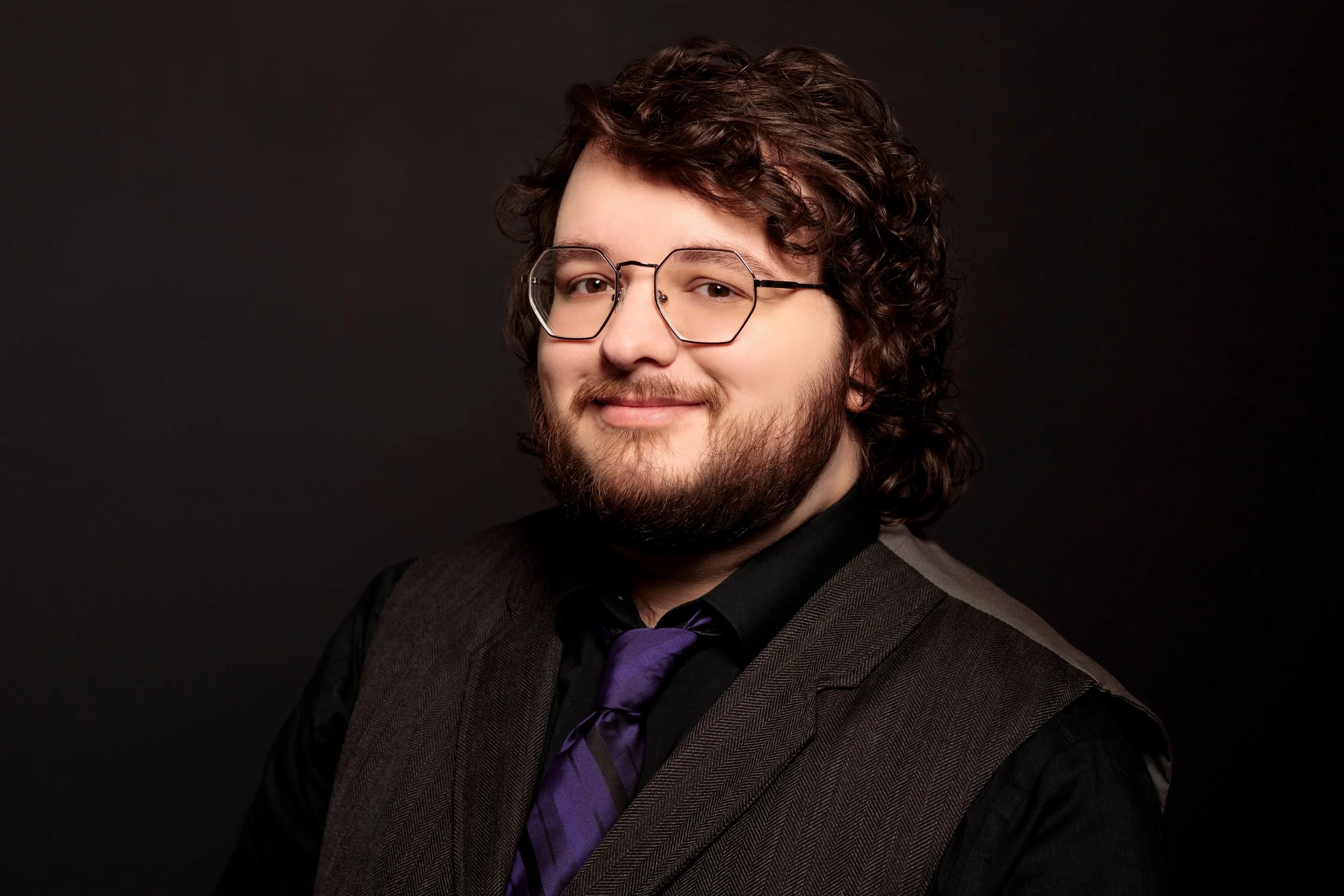A young man with curly brown hair, glasses, and a beard, wearing a black shirt, a purple tie, and a brown vest, smiling against a black background.