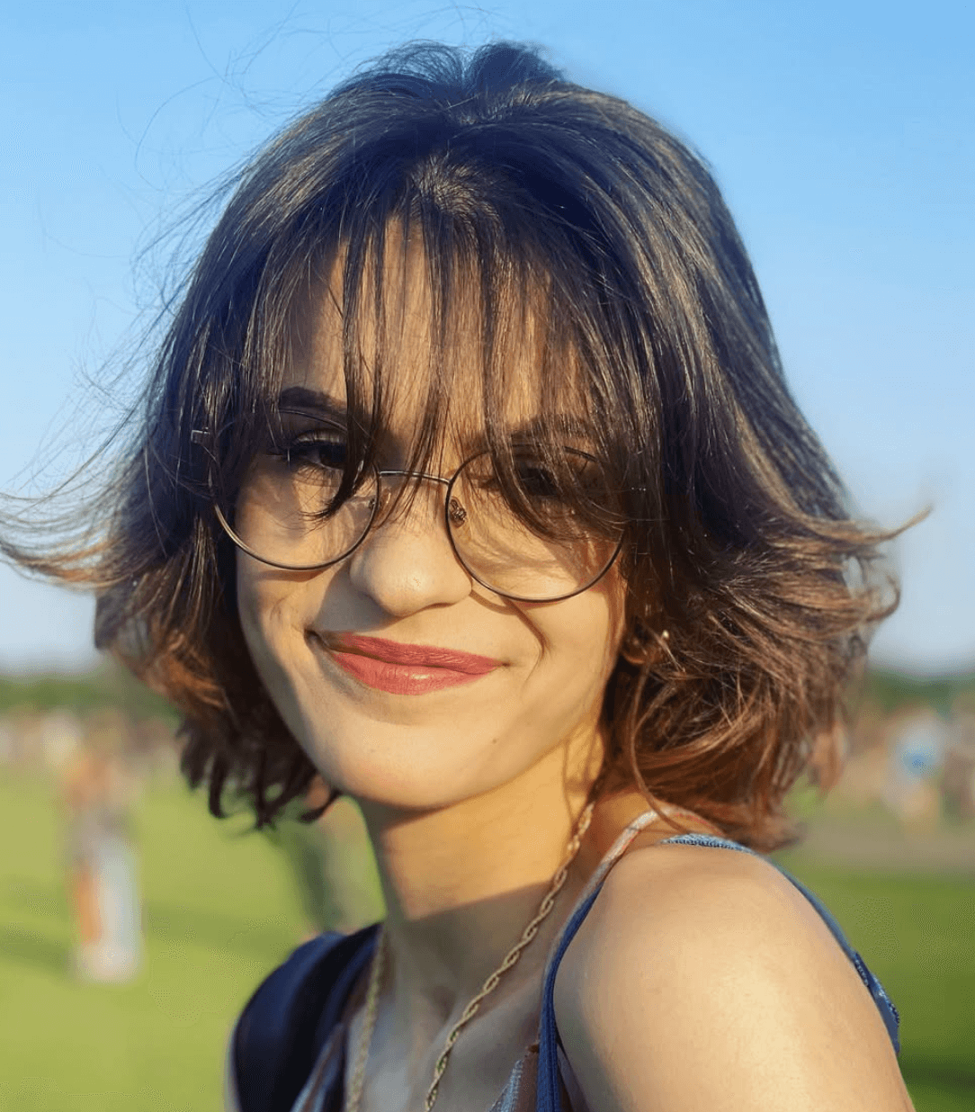 Smiling young person with chin length wavy hair and glasses outdoors in sunlight.