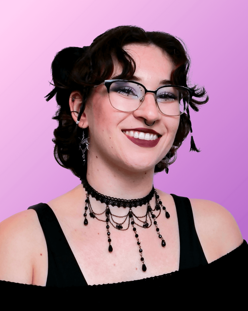 A young person with short curly brown hair, glasses, and multiple earrings, wearing gothic-style black jewelry, smiling against a lilac background.