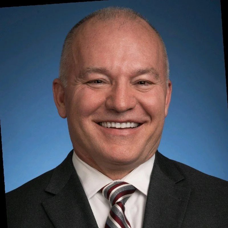 A smiling man in a black suit, white shirt, and striped red, black, and white tie against a blue background.