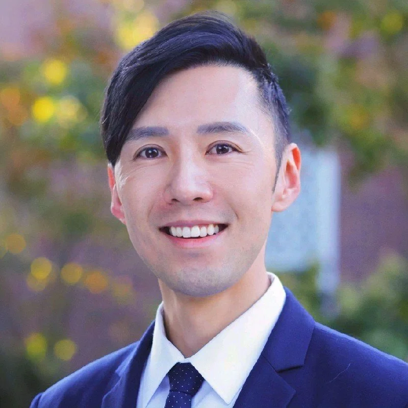 A smiling man in a suit with a blurred outdoor background of trees and sunlight.