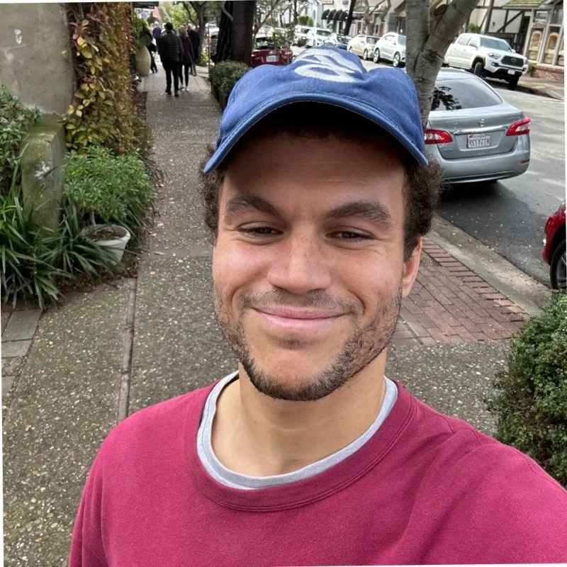 A young man with a beard smiling, wearing a blue cap and a red shirt, taking a selfie on a sidewalk in an urban area with parked cars and trees in the background.