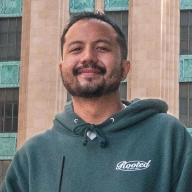 A smiling man with short dark hair and a beard, wearing a dark green hoodie with 'Rooted' logo, standing outside in front of a modern building with large windows and decorative green panels.