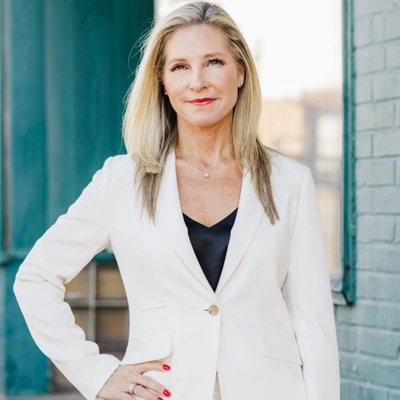 A confident middle-aged woman with long blonde hair, wearing a white blazer over a black top, standing outdoors near teal and brick walls, smiling and looking at the camera.