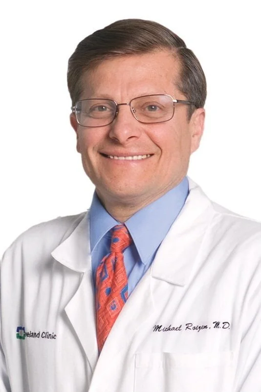 A man wearing glasses in a white medical coat, blue shirt, and patterned tie, smiling at the camera.