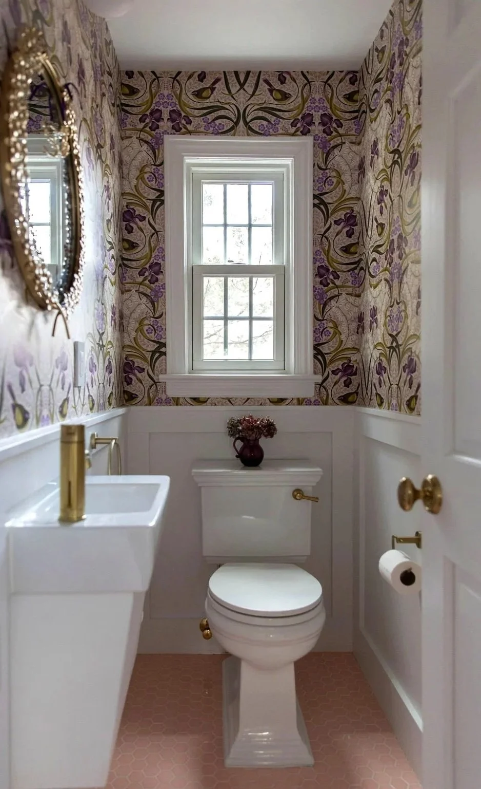 A small bathroom with a toilet, a wall-mounted sink with a gold faucet, a window, floral wallpaper with purple and green patterns, a mirror with a gold frame, and a small vase with pink flowers on top of the toilet tank.