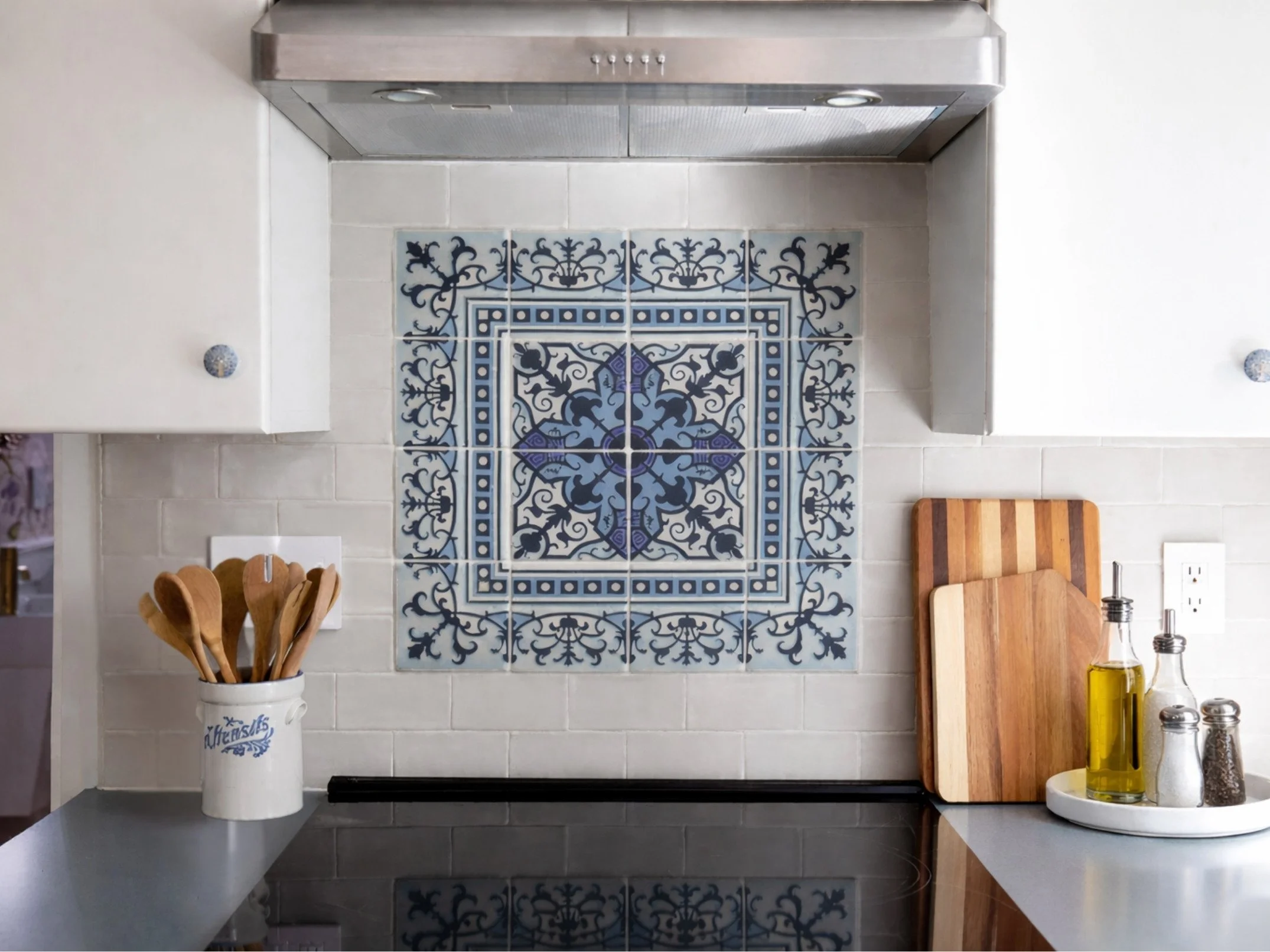 Kitchen designed by Caroline by Design featuring a blue decorative backsplash tile, white cabinetry, gray countertops, brass fixtures, and warm hardwood floors.