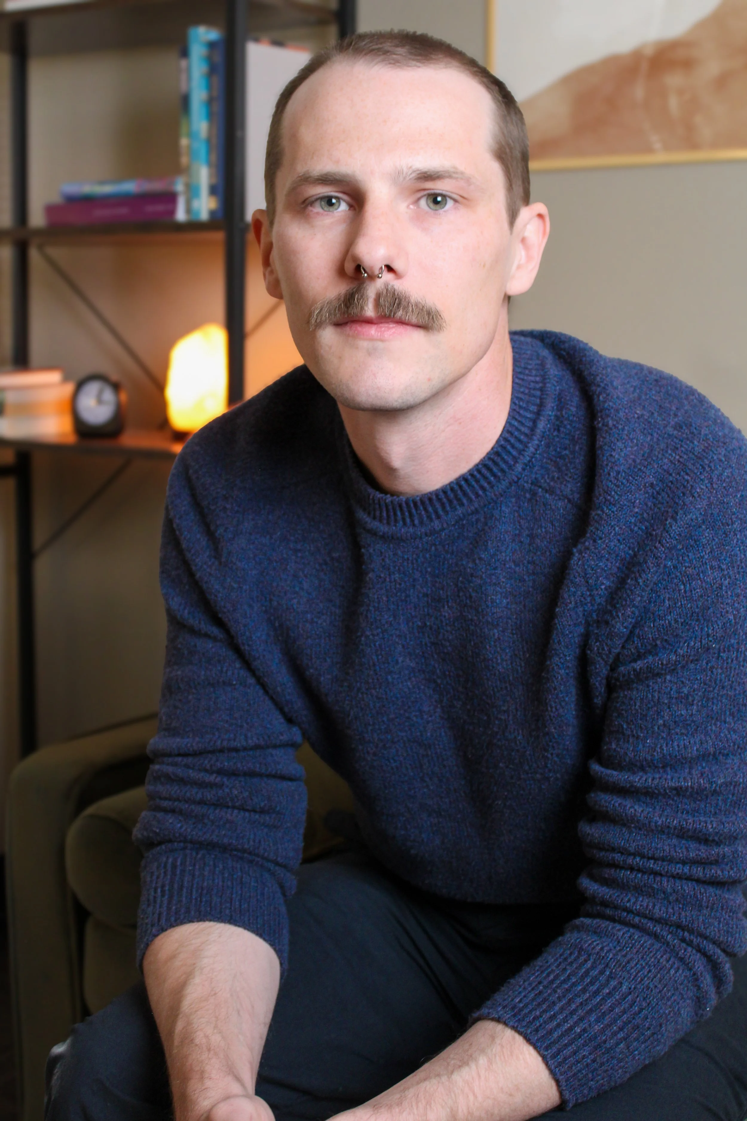 A man with light skin, short brown hair, a mustache, and a septum piercing, wearing a dark blue sweater, sitting in a cozy room with a bookshelf and a lamp in the background.