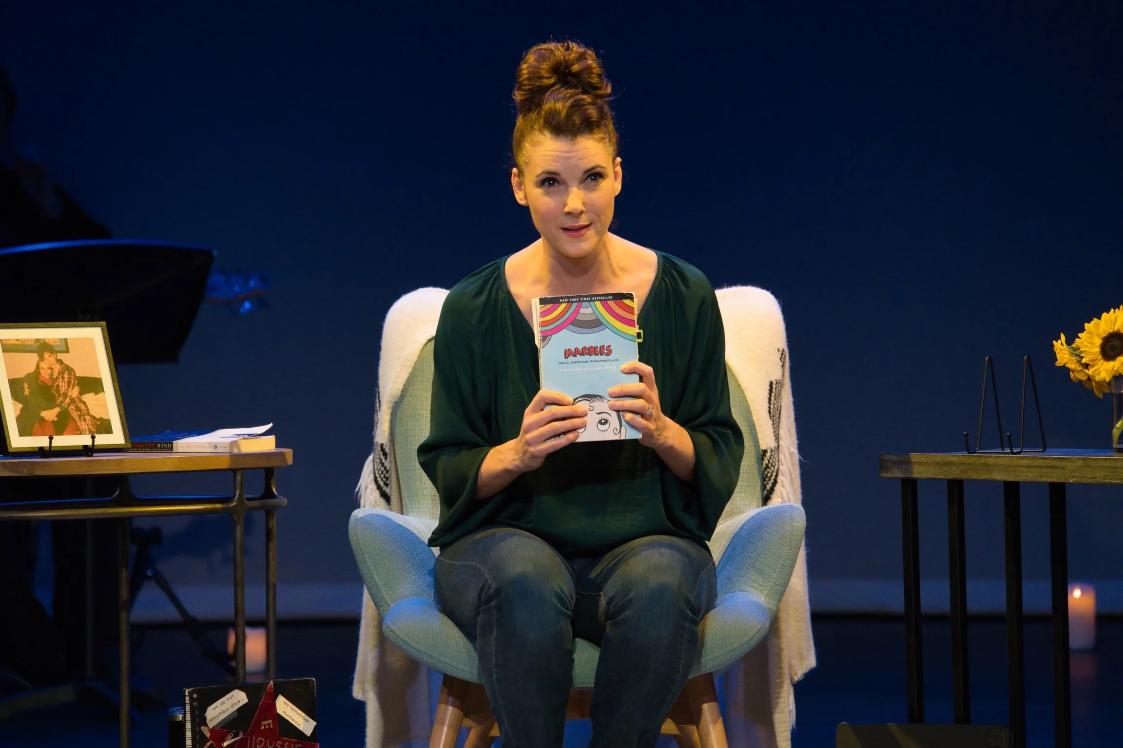 Chryssie Whitehead on stage for In My Own Little Corner, holding a copy of the book 'Marbles' and looking at the camera.