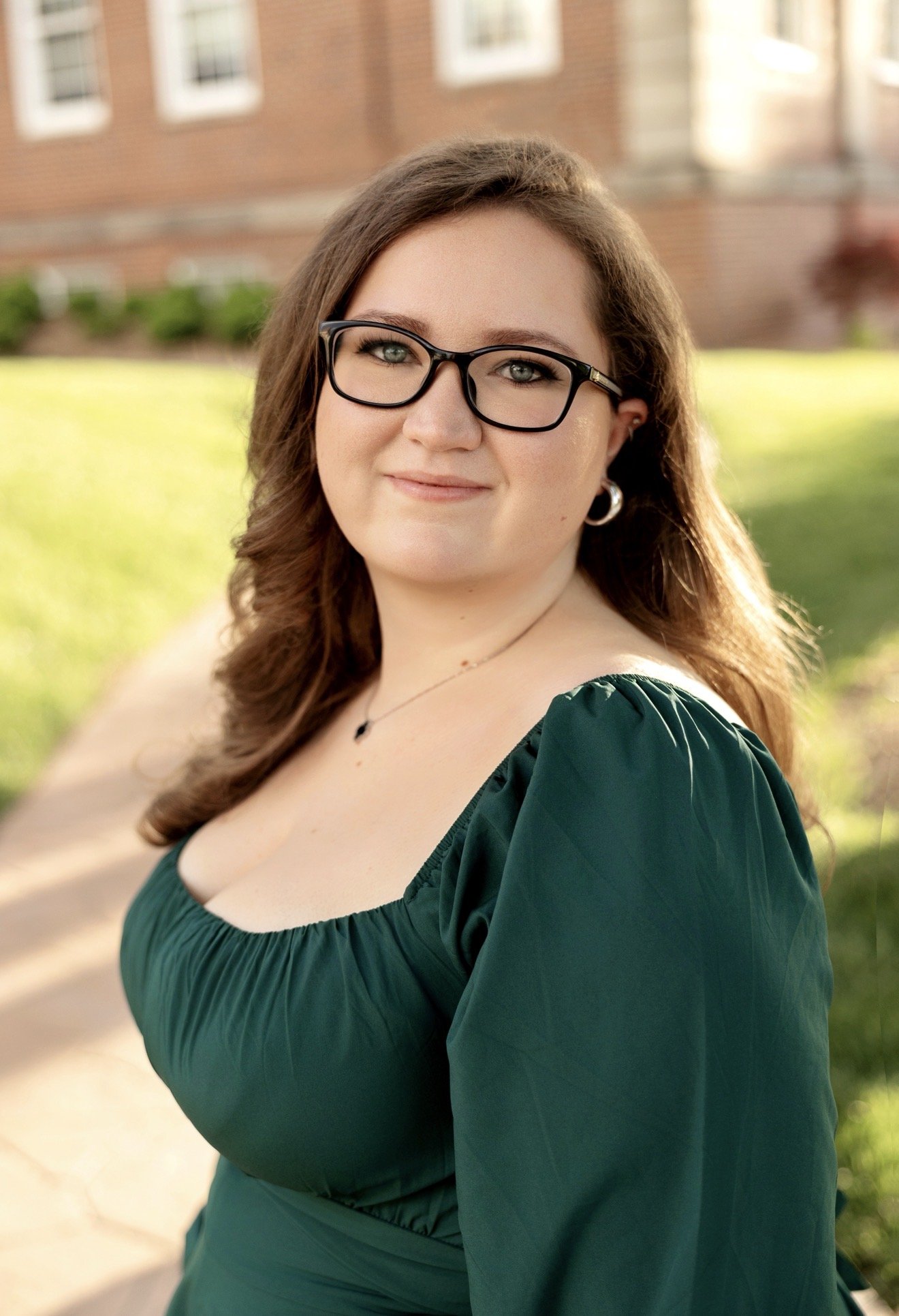 Headshot of woman outside with brown hair, black glasses, and green dress