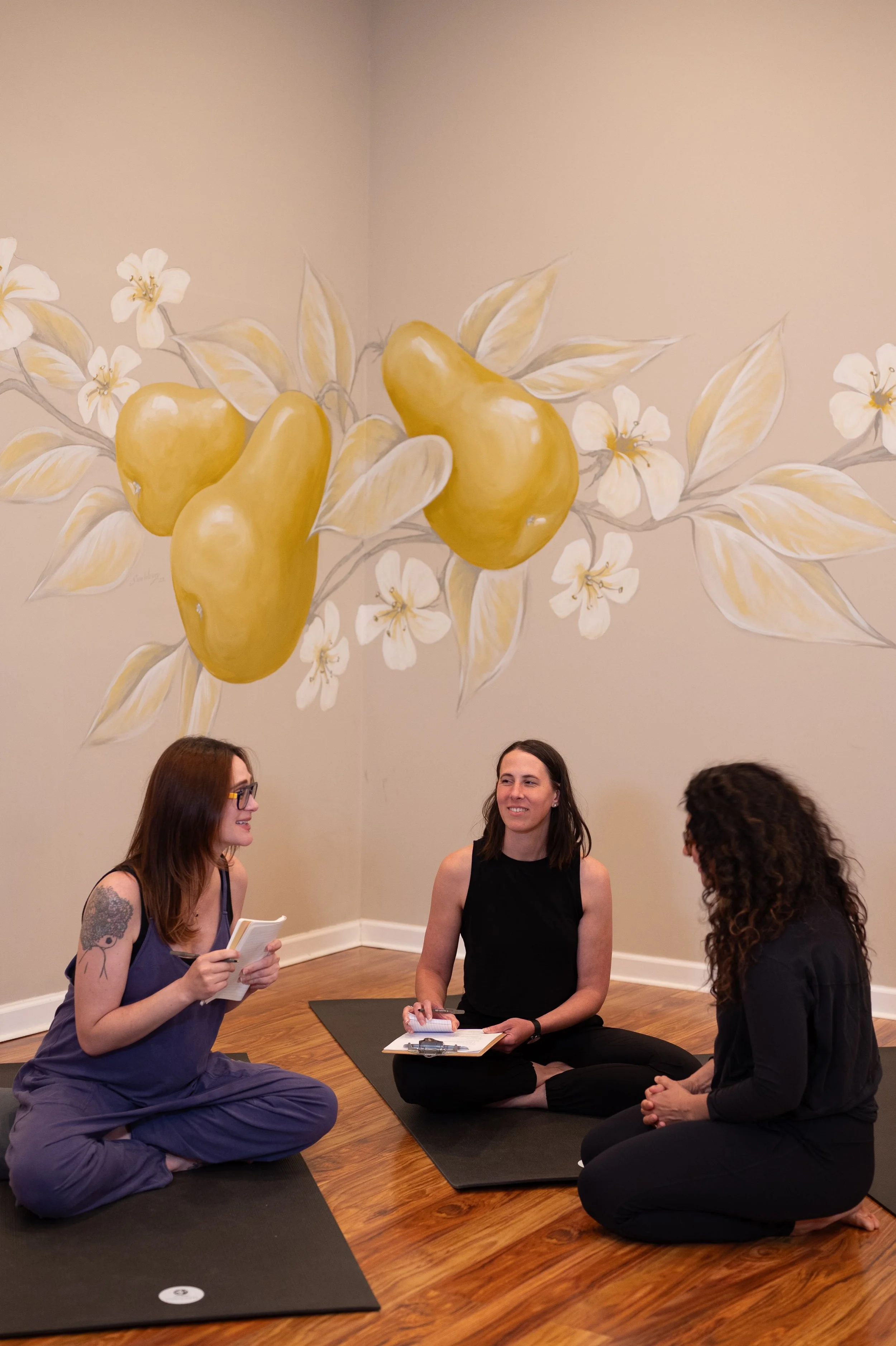 Three female yoga instructors chatting on mats in front of pear mural
