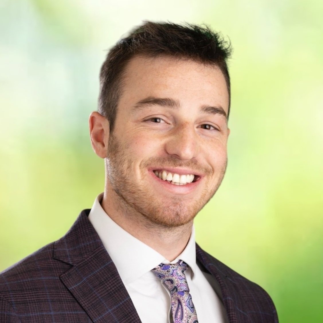 A smiling young man with short dark hair, wearing a dark checkered suit, white shirt, and a patterned tie, standing against a blurred green background.