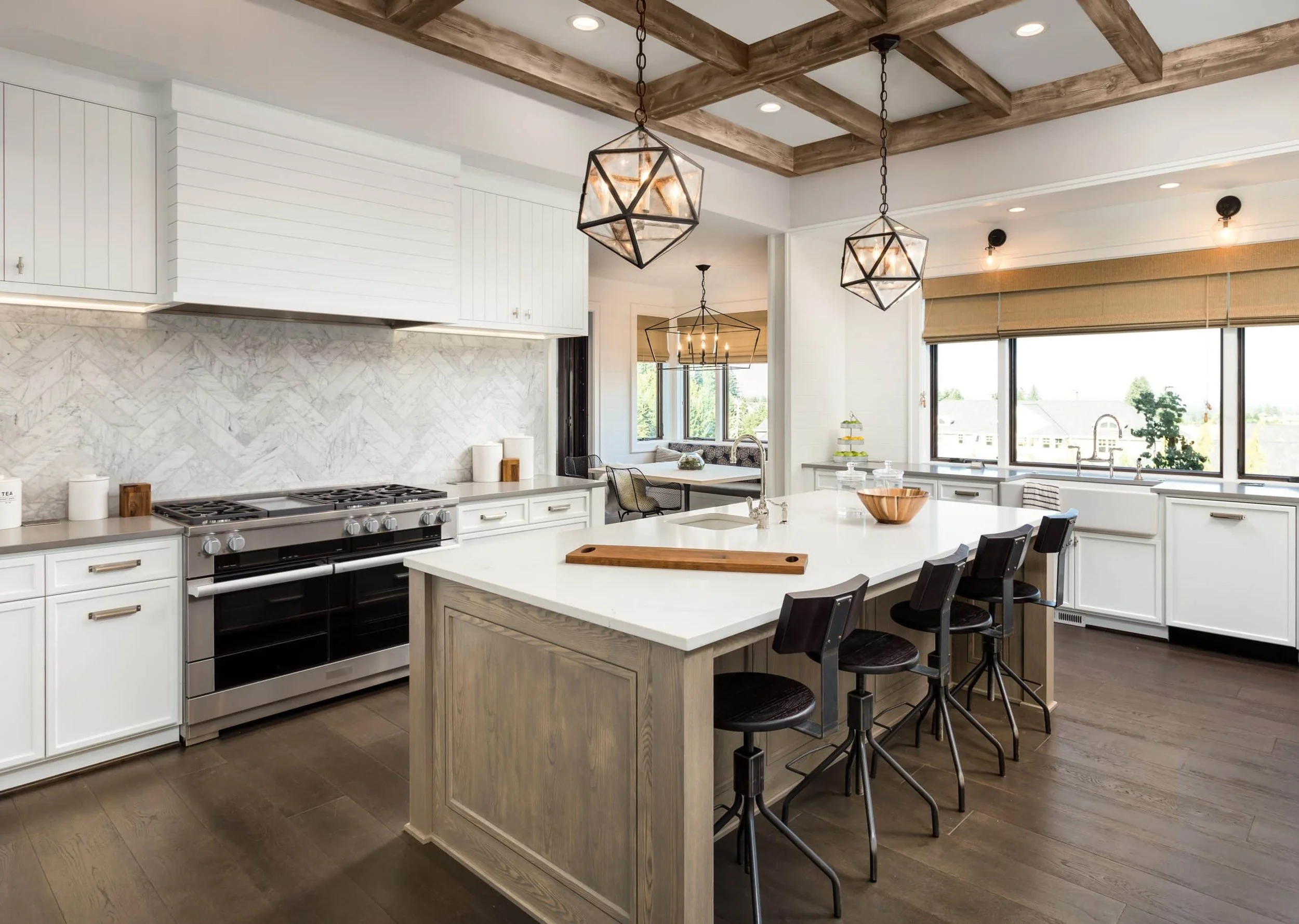 Modern kitchen with white cabinets, marble backsplash, stainless steel stove, central island with white countertop, dark wood bar stools, large windows, and wooden ceiling beams.