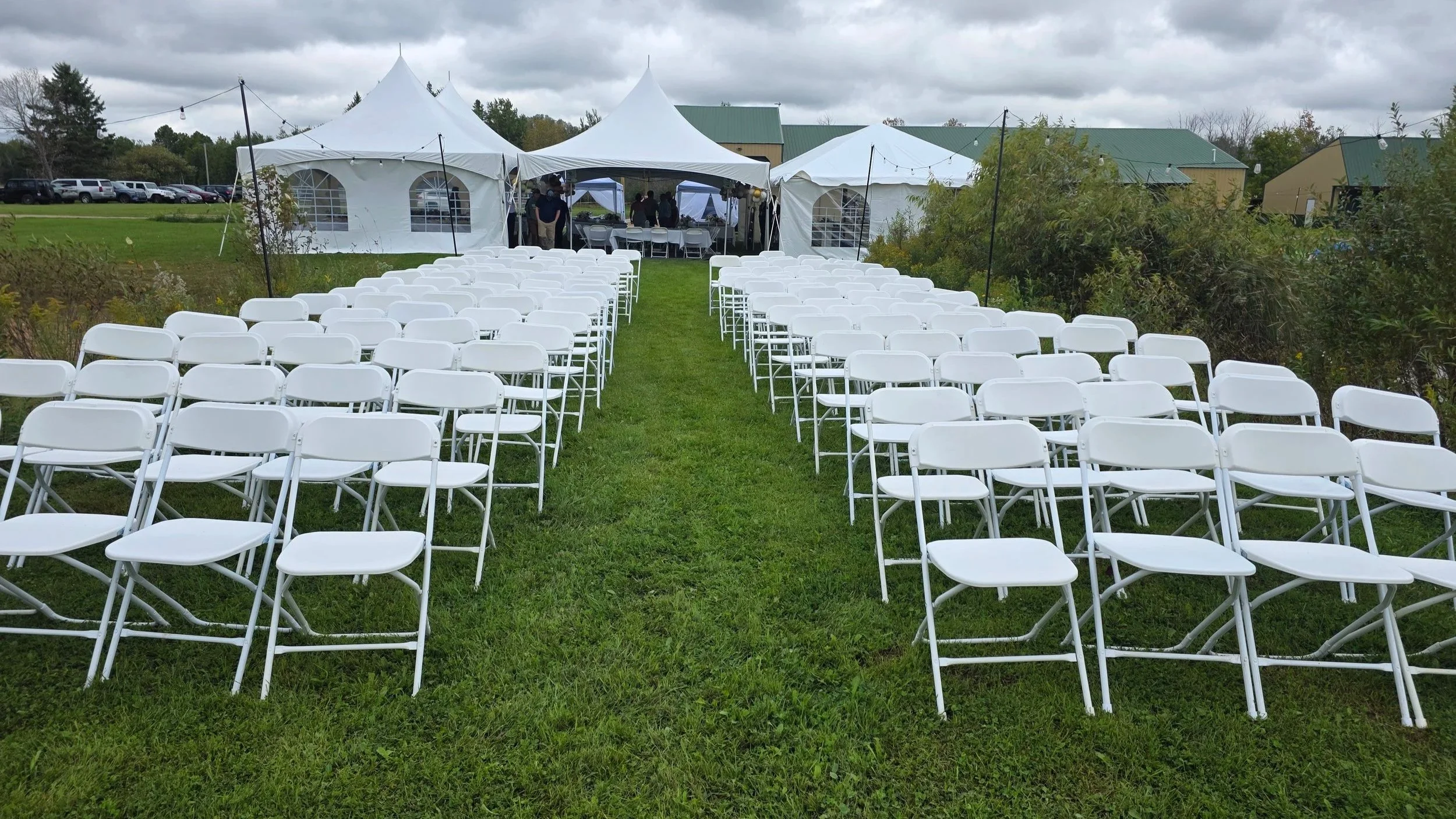 Outdoor wedding ceremony setup with multiple white folding chairs arranged in rows on a grassy lawn, facing white tents with peaked roofs and windows, under a cloudy sky.