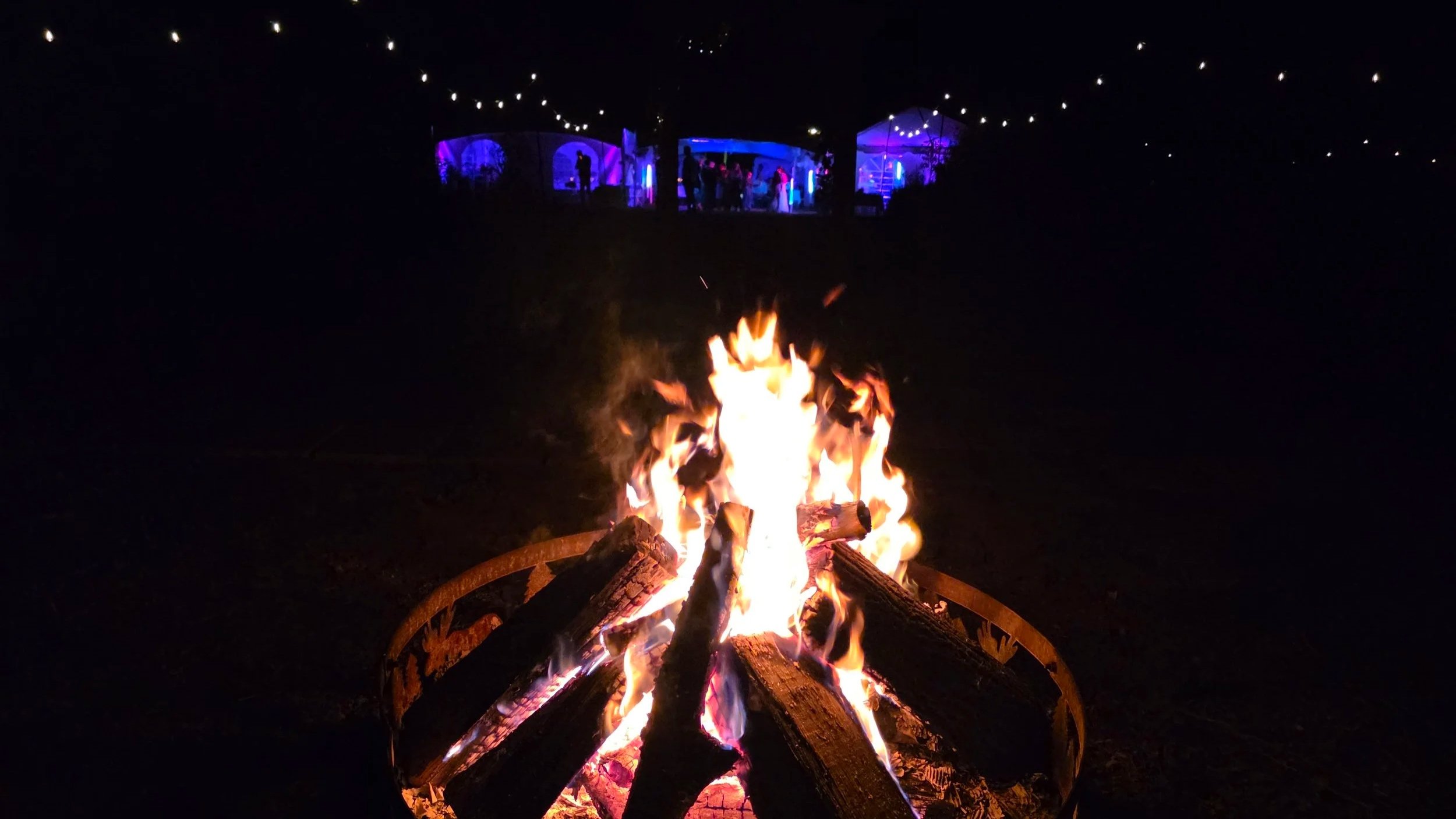 A bonfire burning with logs in the foreground and a nighttime outdoor gathering or party with string lights and tents illuminated in blue and purple in the background.