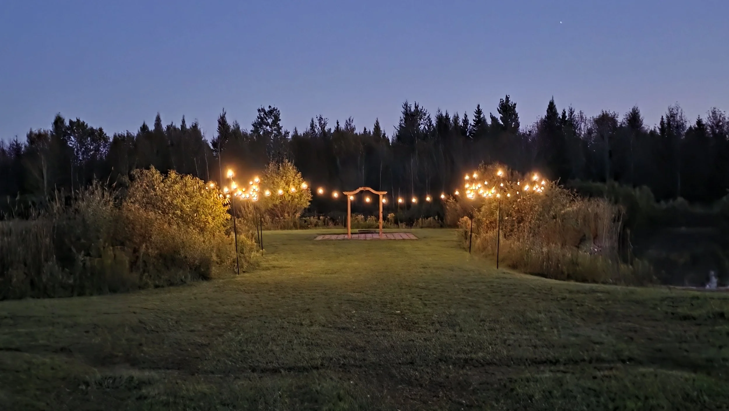 Outdoor wedding ceremony setup with multiple white folding chairs arranged in rows on a grassy lawn, facing white tents with peaked roofs and windows, under a cloudy sky.