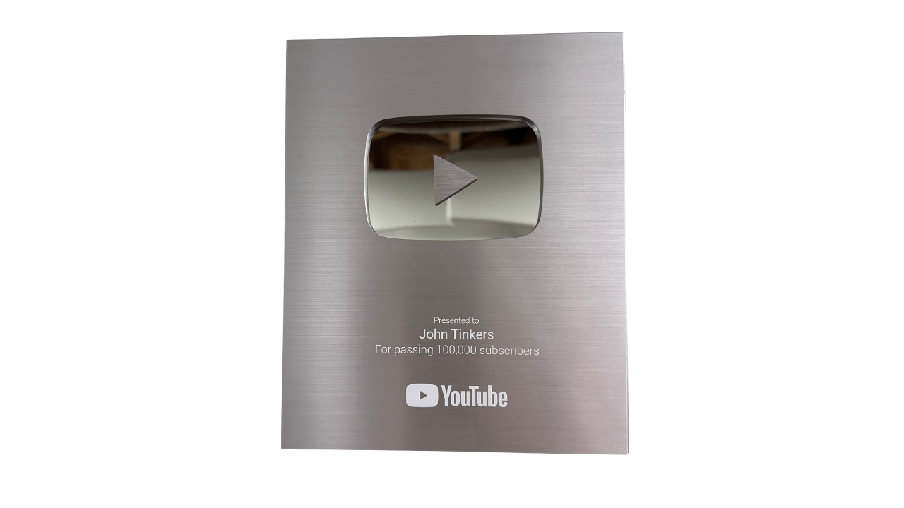 Silver YouTube silver play button award plaque with an engraved play icon and text recognizing John Tinkers for passing 100,000 subscribers.