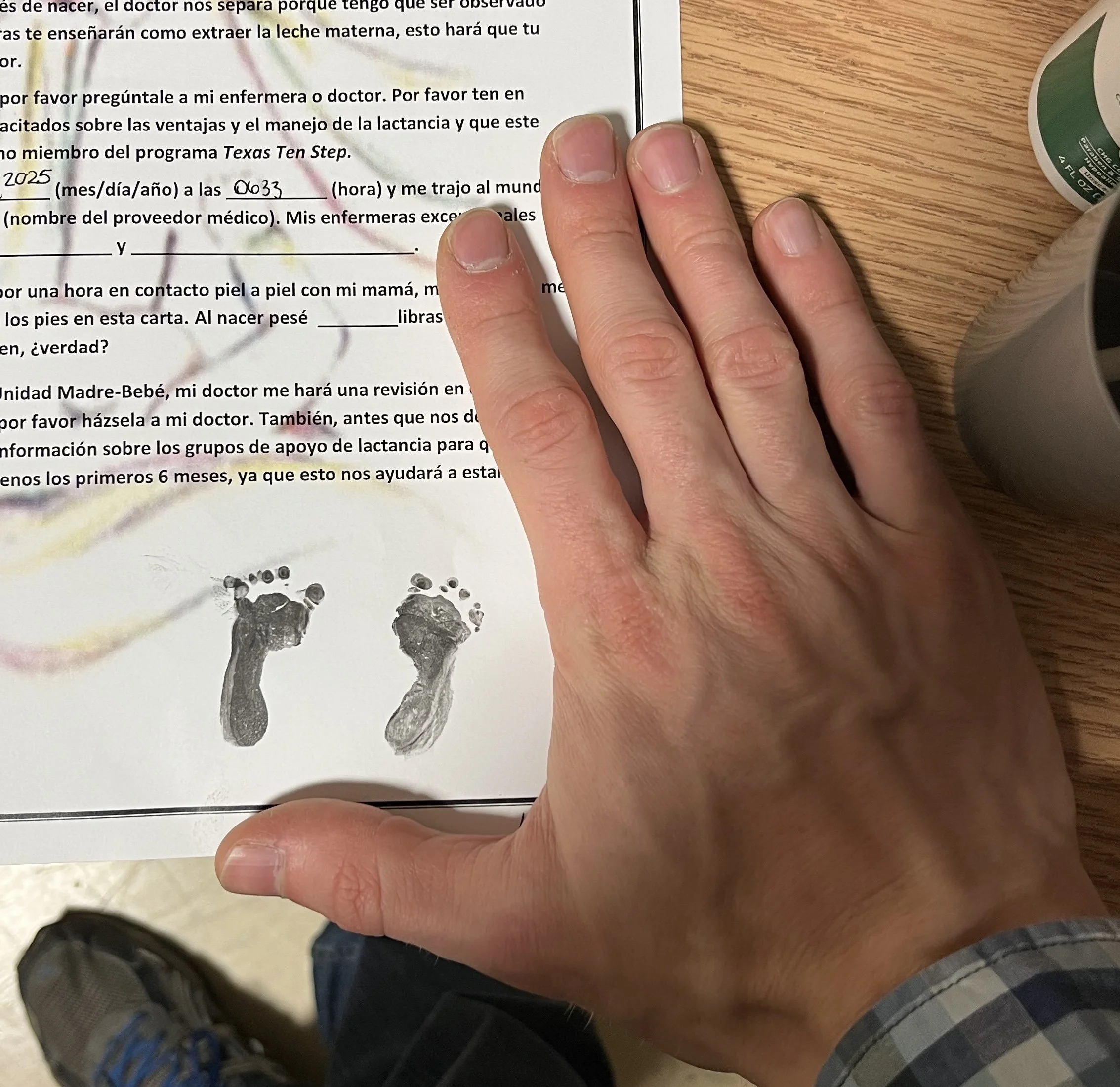 A person's left hand resting on a paper with black text, showing fingerprints near the bottom. The person is wearing a plaid shirt sleeve, and there is a wooden surface and a green container partially visible nearby. Feet wearing blue sneakers are visible at the bottom of the image.