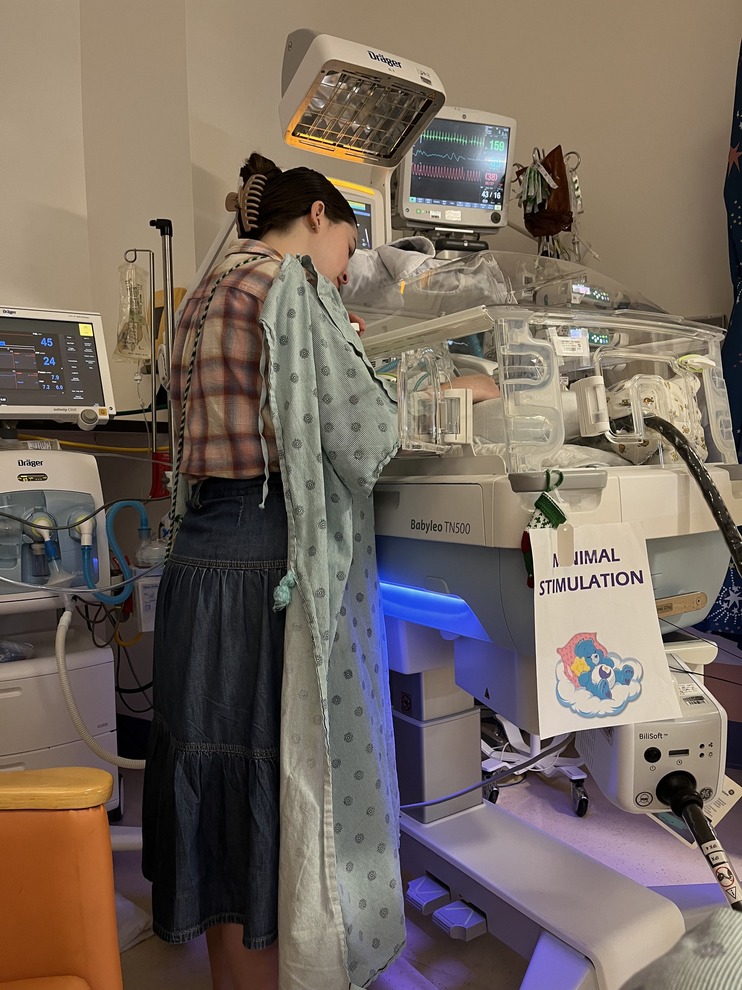 A woman is looking in a neonatal incubator in a hospital, connected to multiple medical monitors and equipment, with an IV attached nearby. There is a sign that reads 'Minimal Stimulation' with a picture of a blue unicorn on a cloud.