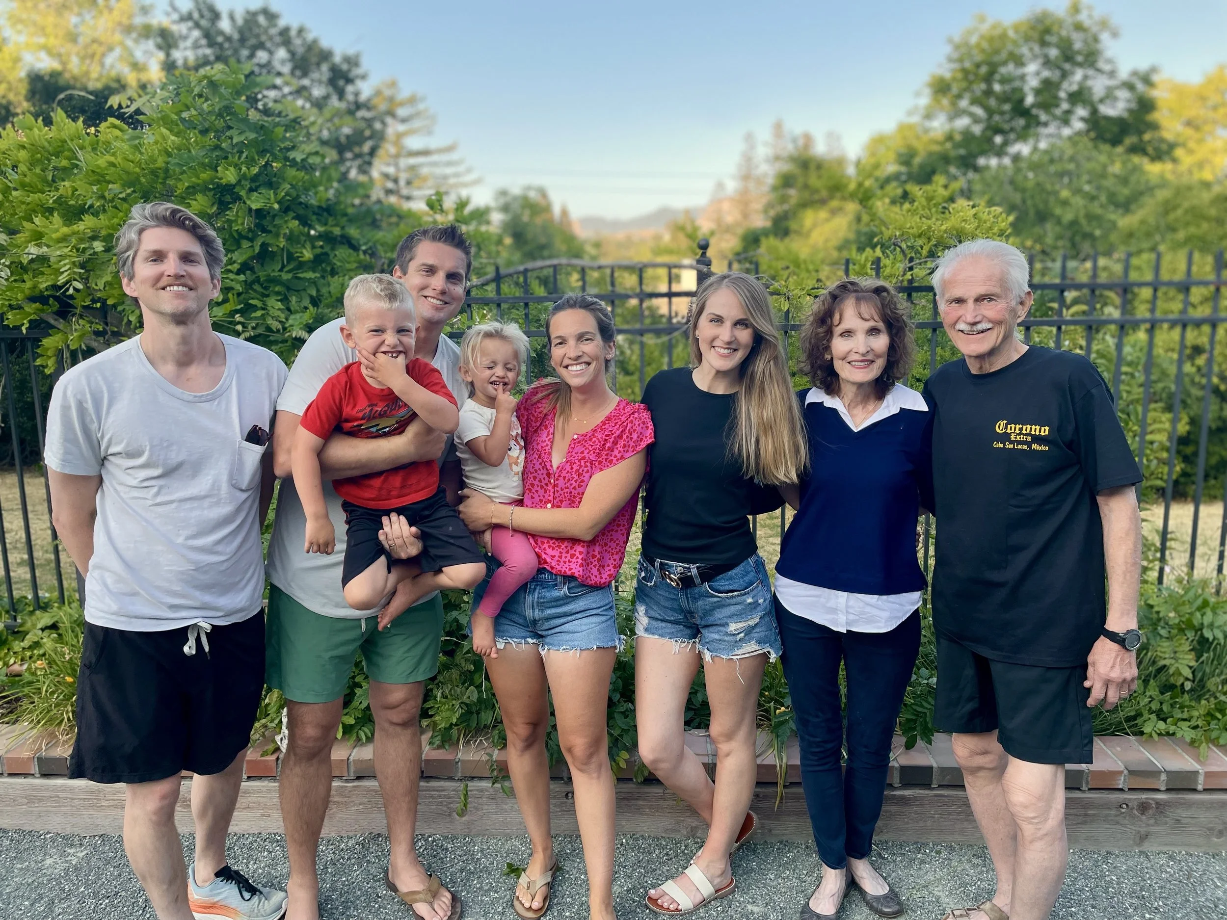 A large group of eight people, including children and adults, standing outdoors in front of a black fence and lush green trees, smiling for a group photo on a clear day.