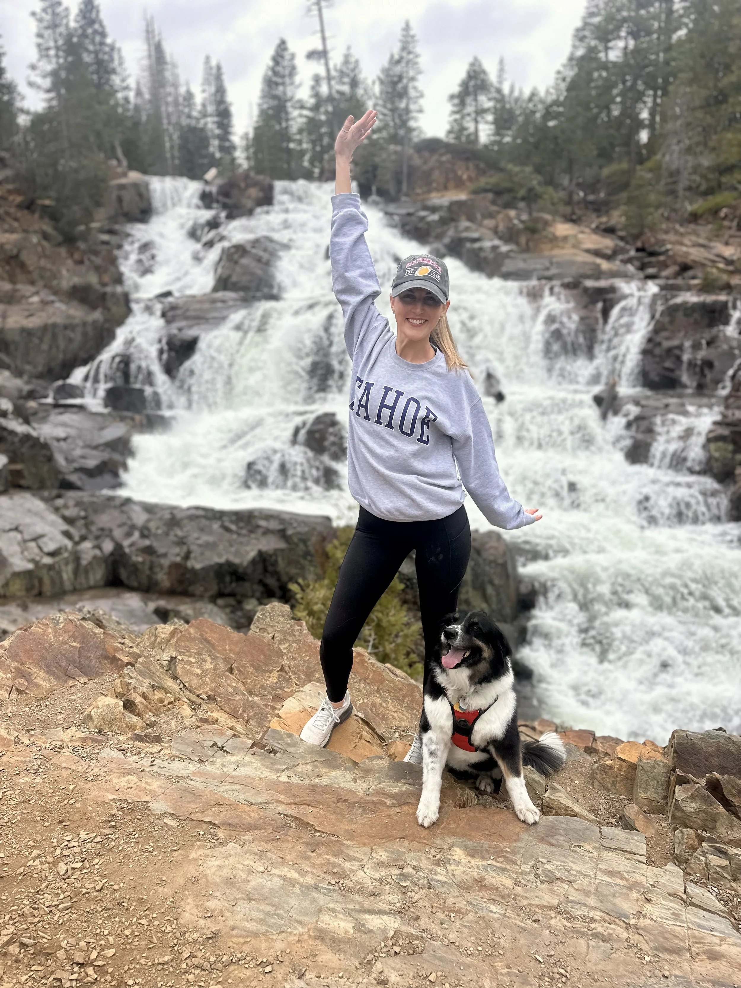 A woman in a gray sweatshirt and black leggings standing on a rocky ledge with a black and white dog in front of a waterfall surrounded by pine trees.