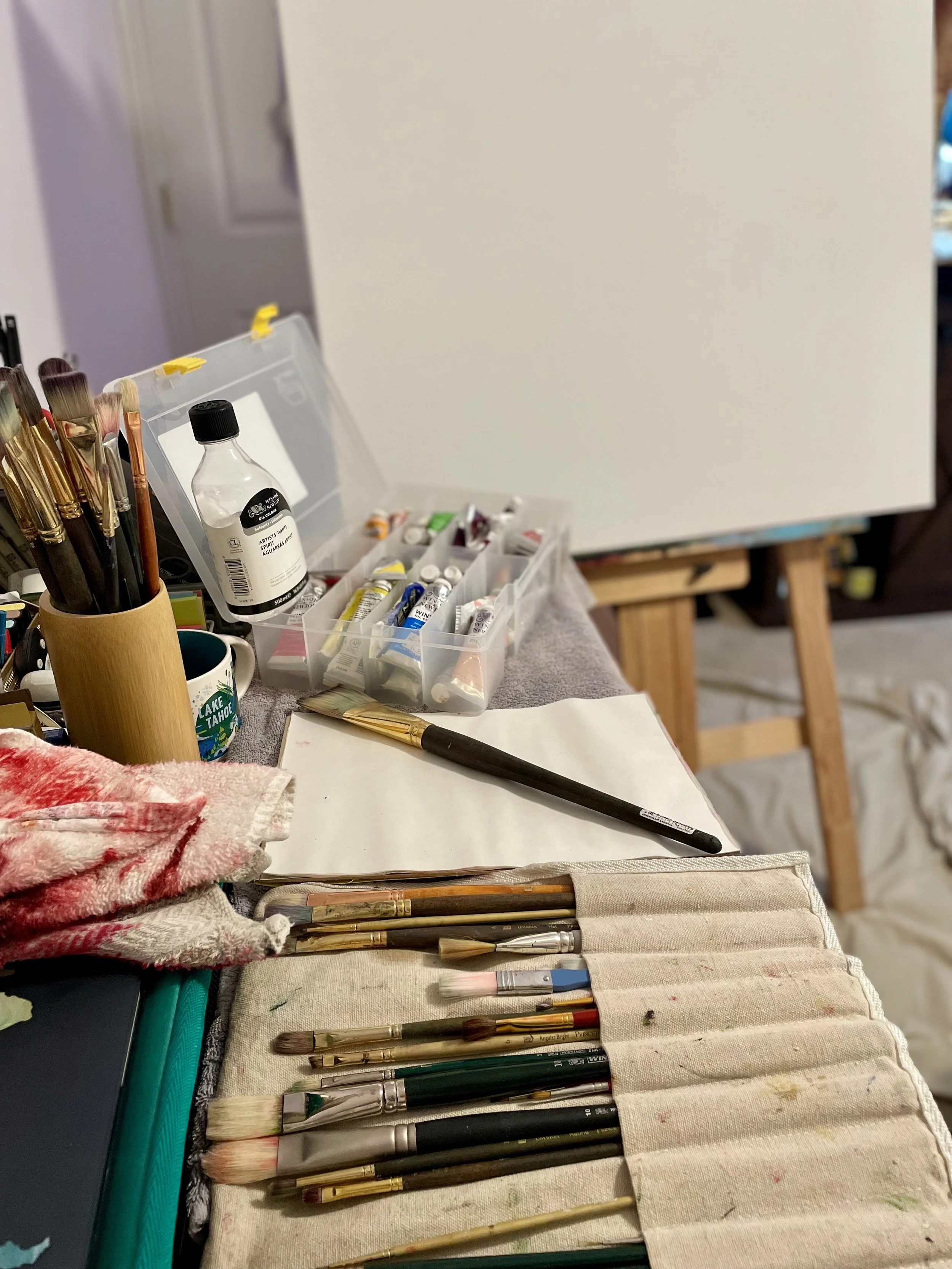 An artist's workspace with various paint brushes, tubes of paint, a palette, a water container, and an easel with a blank canvas.