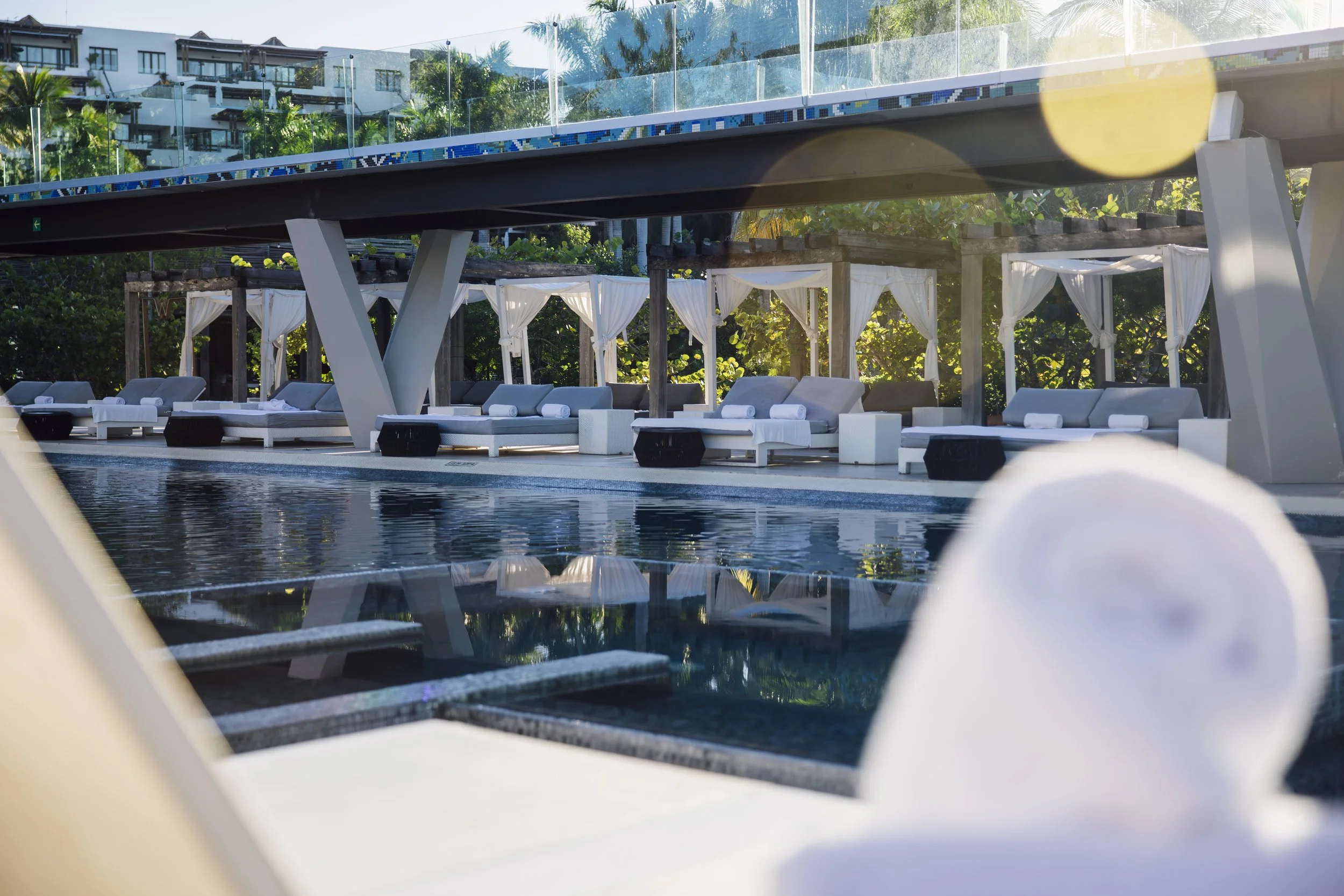 A luxurious poolside scene with white lounge chairs and cabanas draped with white curtains, shaded by wooden beams, under a canopy of green trees, with a modern multi-story building in the background, captured in a sunny setting.