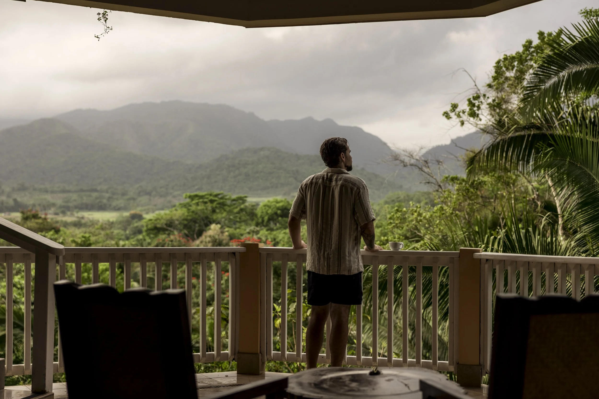 A man stands on a balcony, looking out over a lush green landscape with mountains in the background on a cloudy day. There is a cup on the railing and some chairs in the foreground.