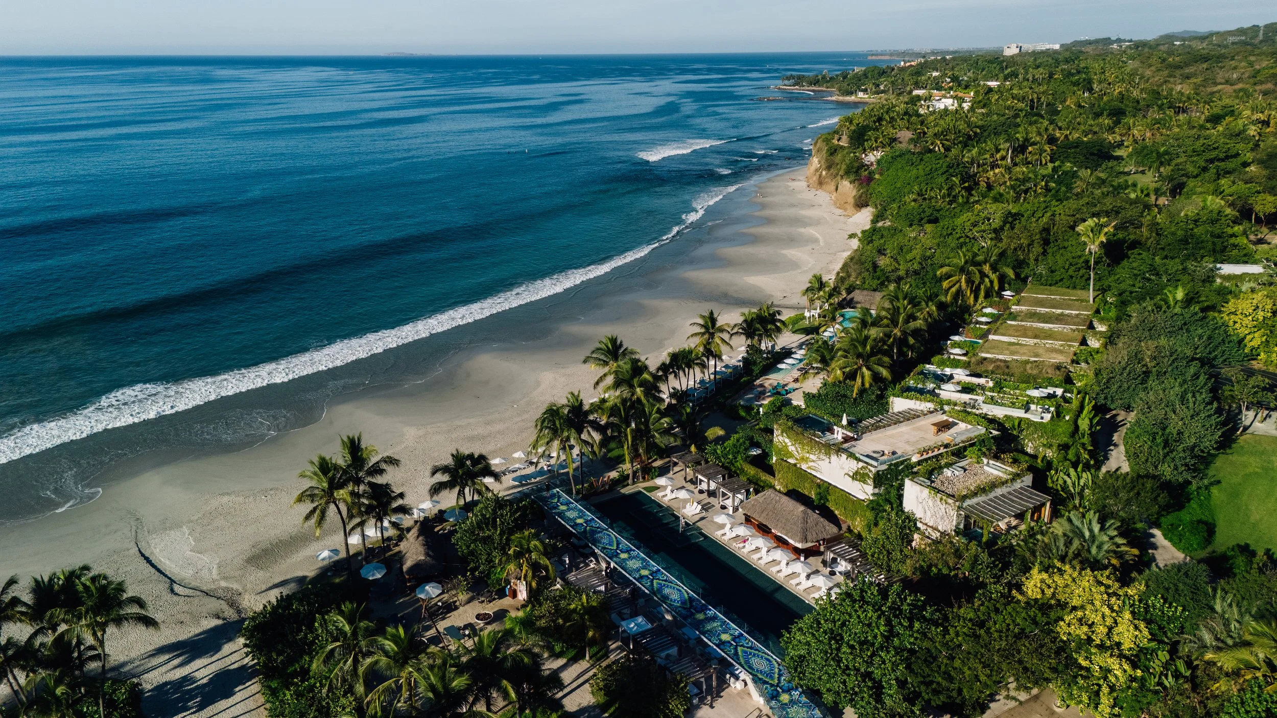 Aerial view of a tropical beach with white sand, rows of palm trees, and resort structures, overlooking the clear blue ocean with gentle waves.