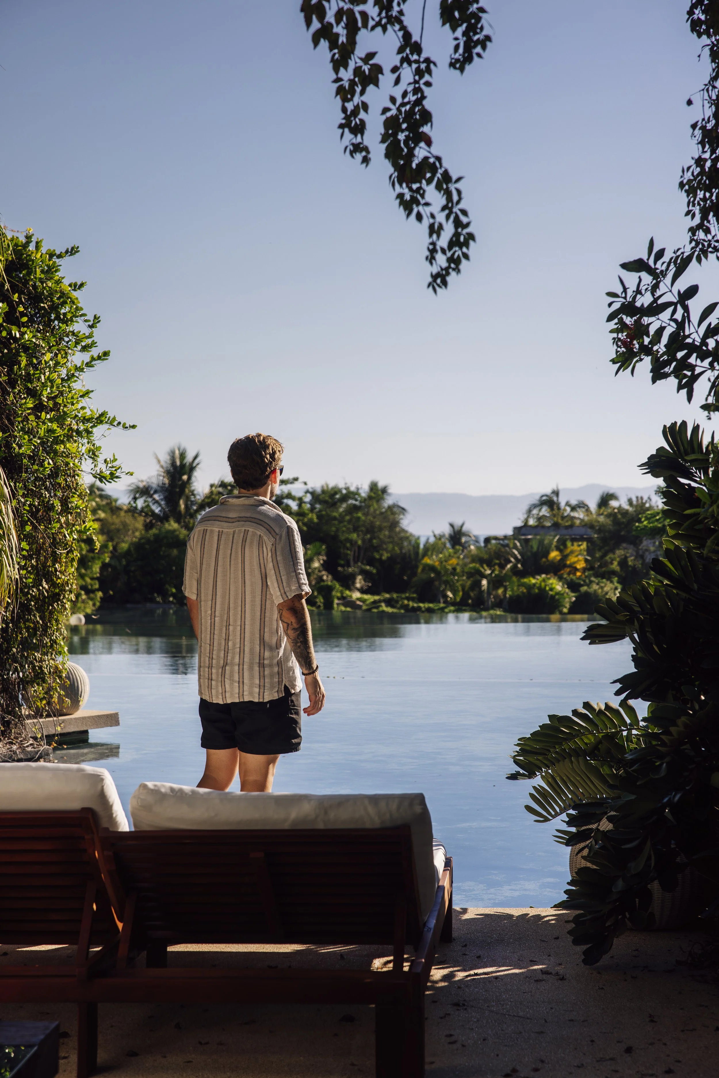 A man with curly hair, wearing sunglasses, a striped short-sleeve shirt, and black shorts, stands by a pool overlooking a body of water with trees and mountains in the background. The scene appears to be in a warm climate during daytime.