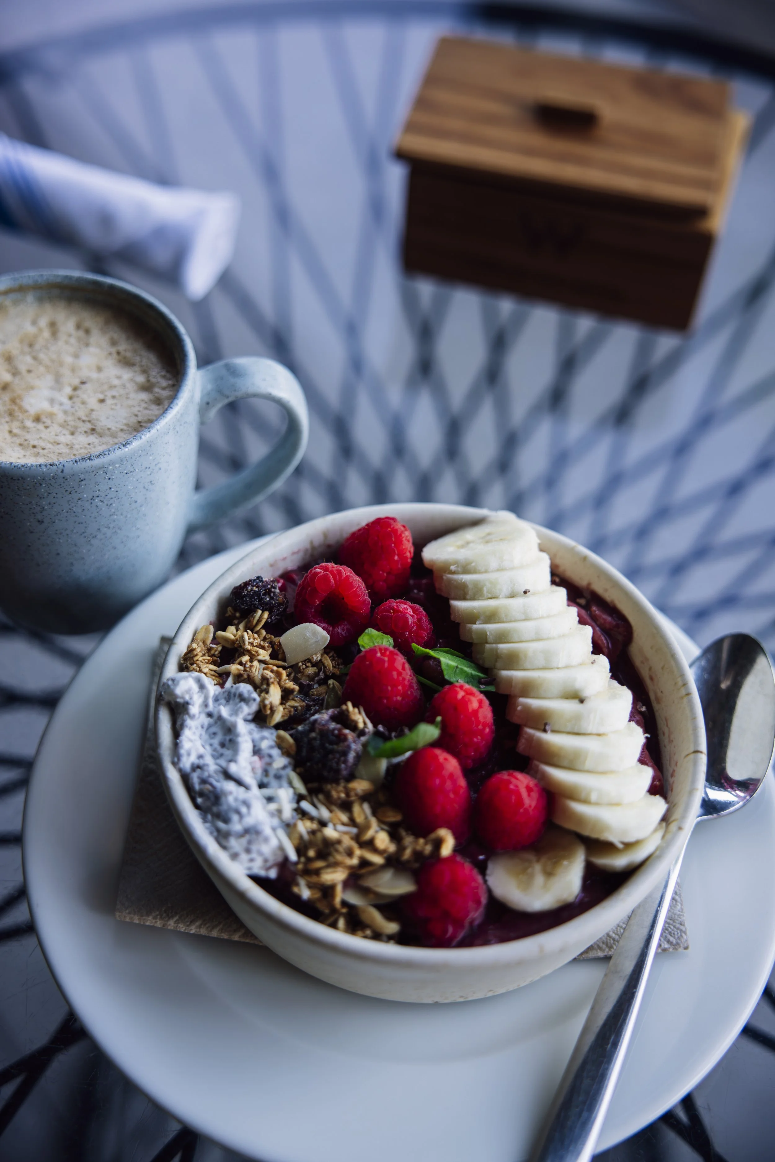 A bowl of yogurt topped with granola, banana slices, and raspberries, served on a white plate with a spoon, alongside a mug of coffee and a wooden box on a table.