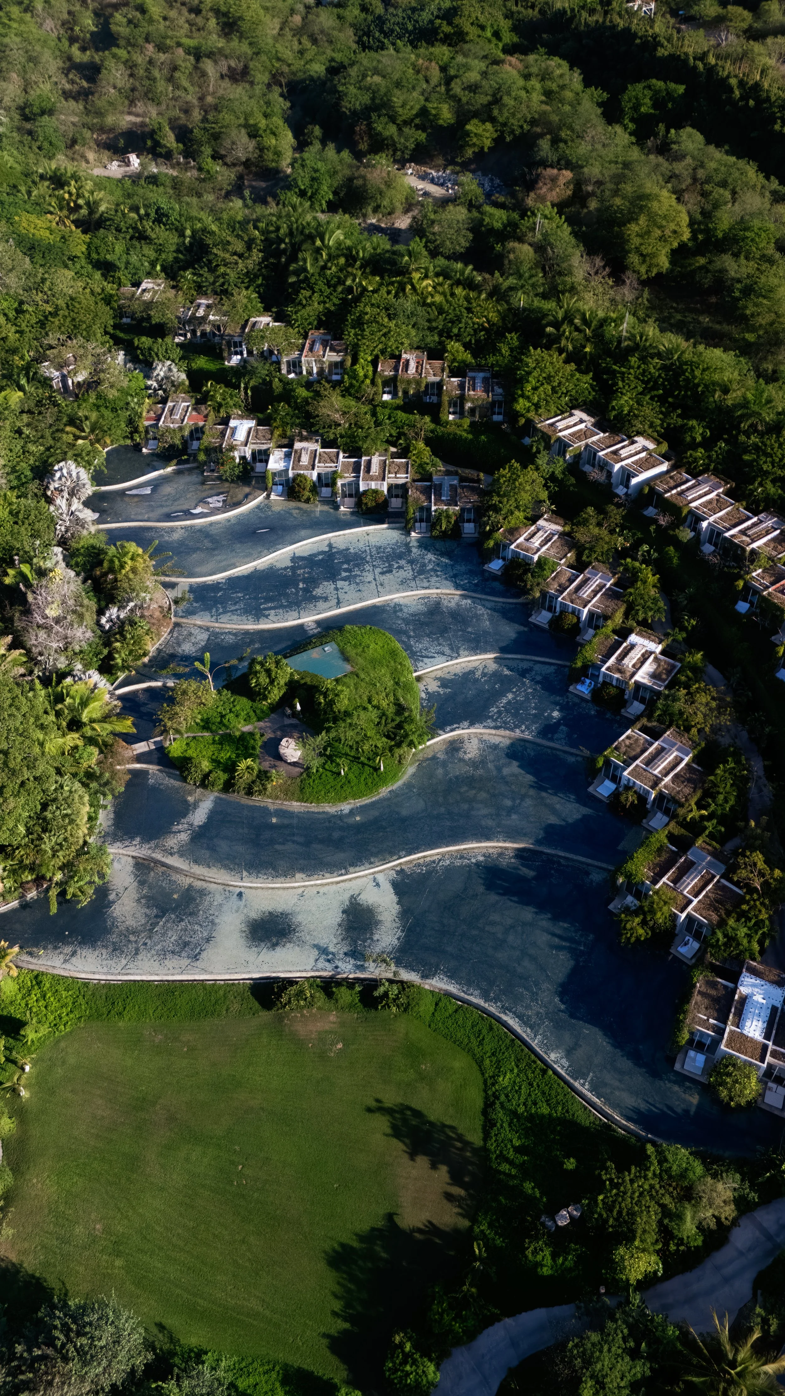 Aerial view of a resort with multiple white buildings, winding pathways, and a large saltwater swimming pool surrounded by lush greenery and trees.