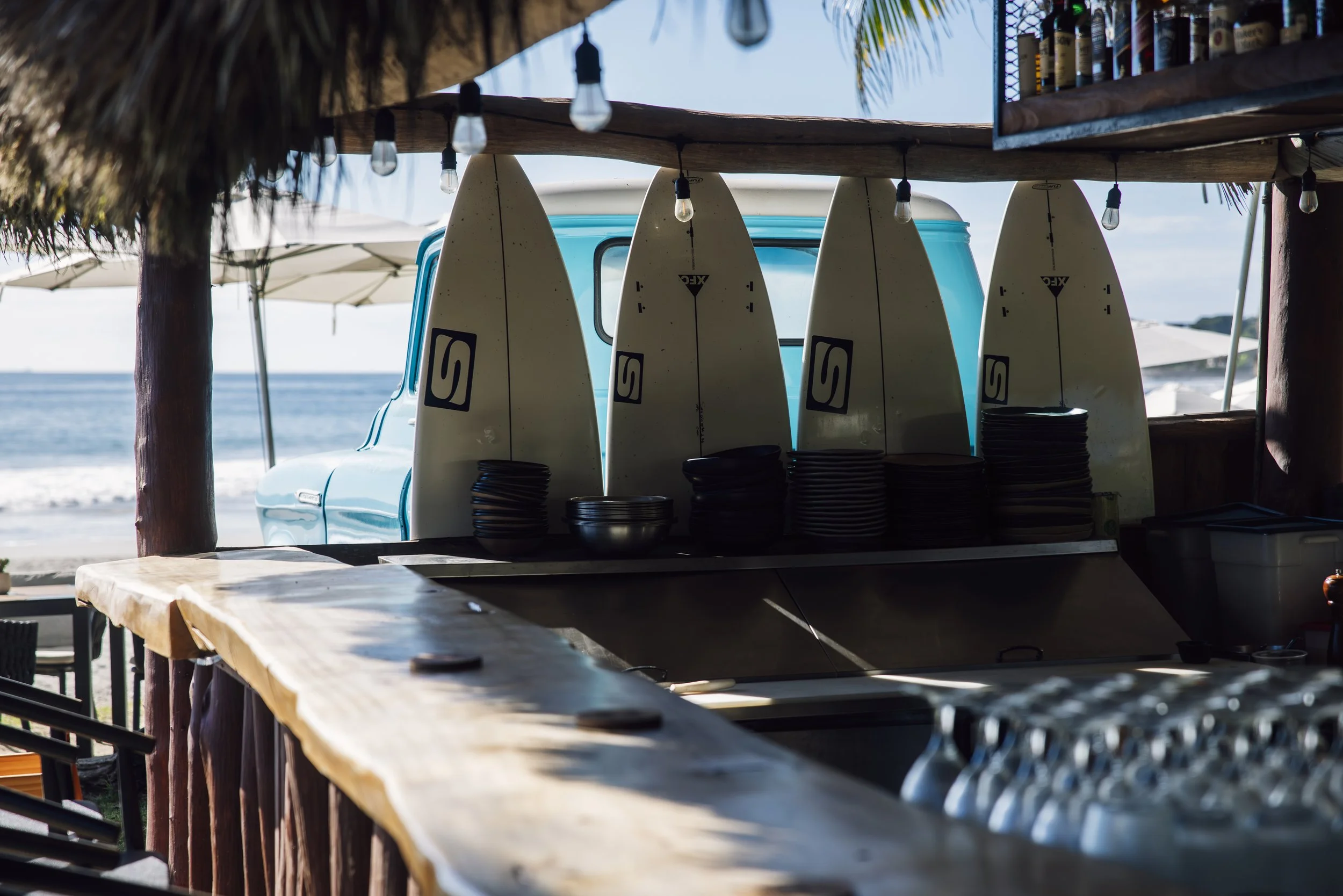 Beachside bar with surfboards, bottles, and plates, overlooking the ocean, with beach umbrella and thatched roofing