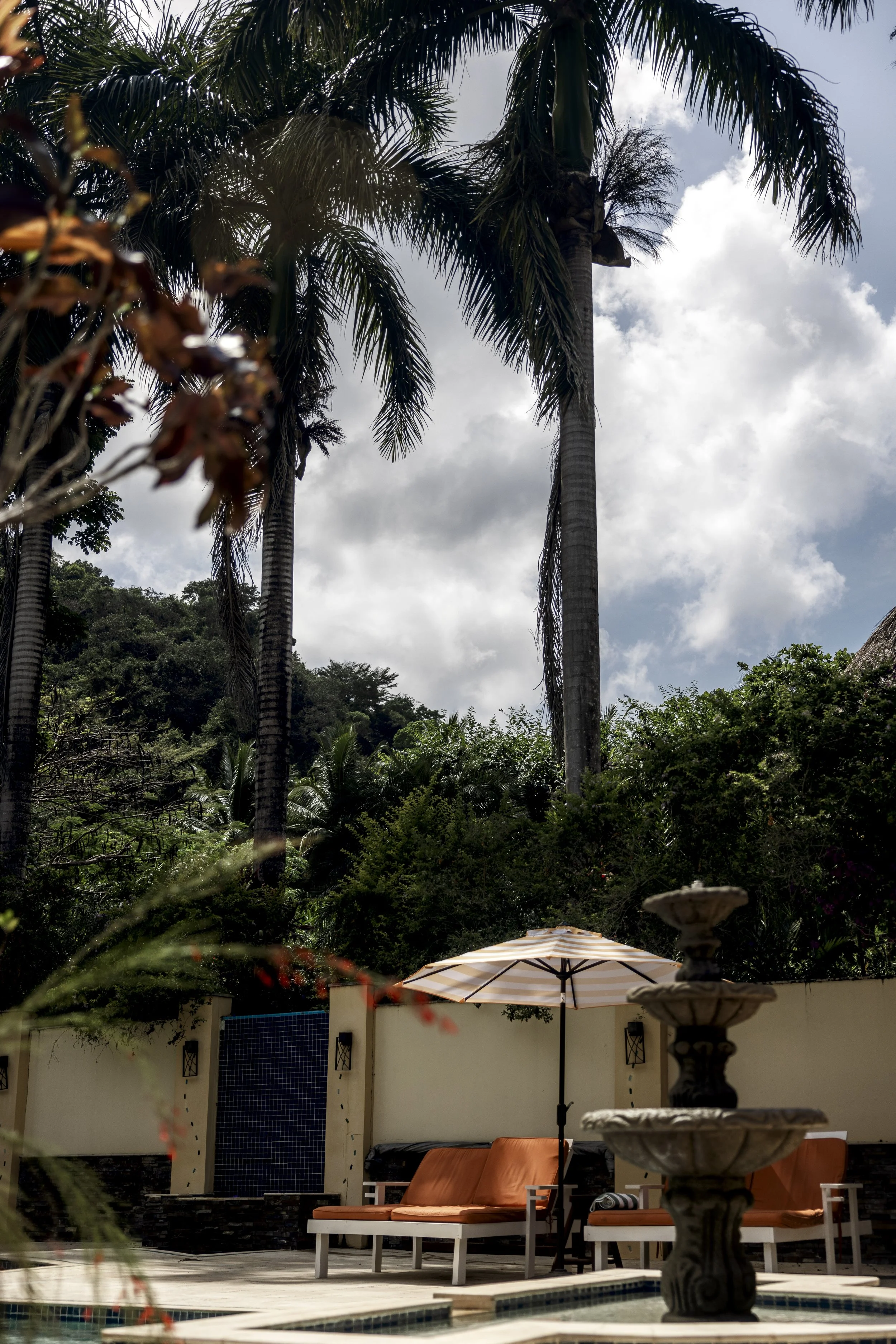 A backyard patio with orange cushioned lounge chairs, a large striped umbrella, a fountain, and lush tropical trees, including tall palm trees under a partly cloudy sky.