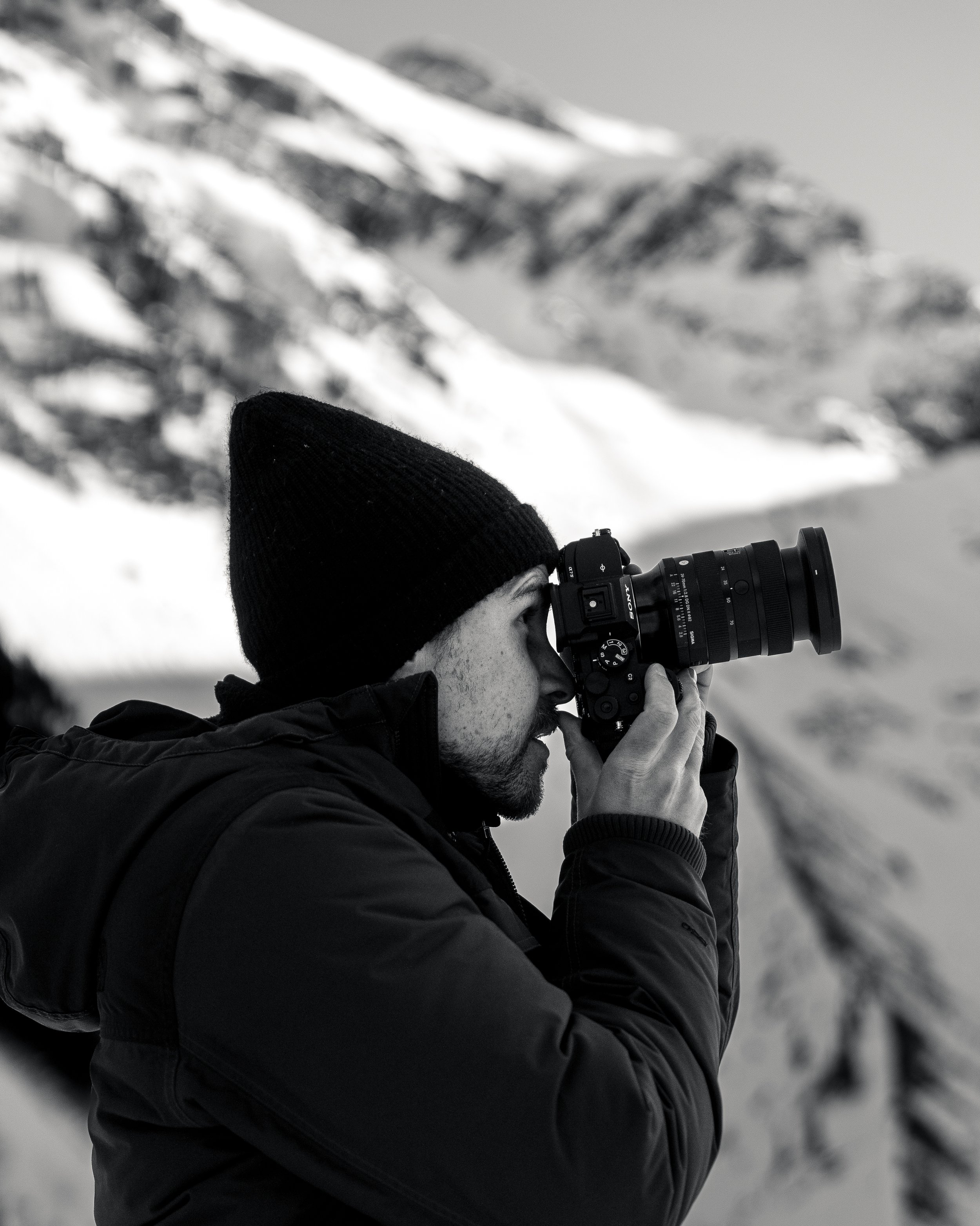 A man wearing a black beanie and black jacket is looking through a camera with a long zoom lens in a snowy outdoor setting.