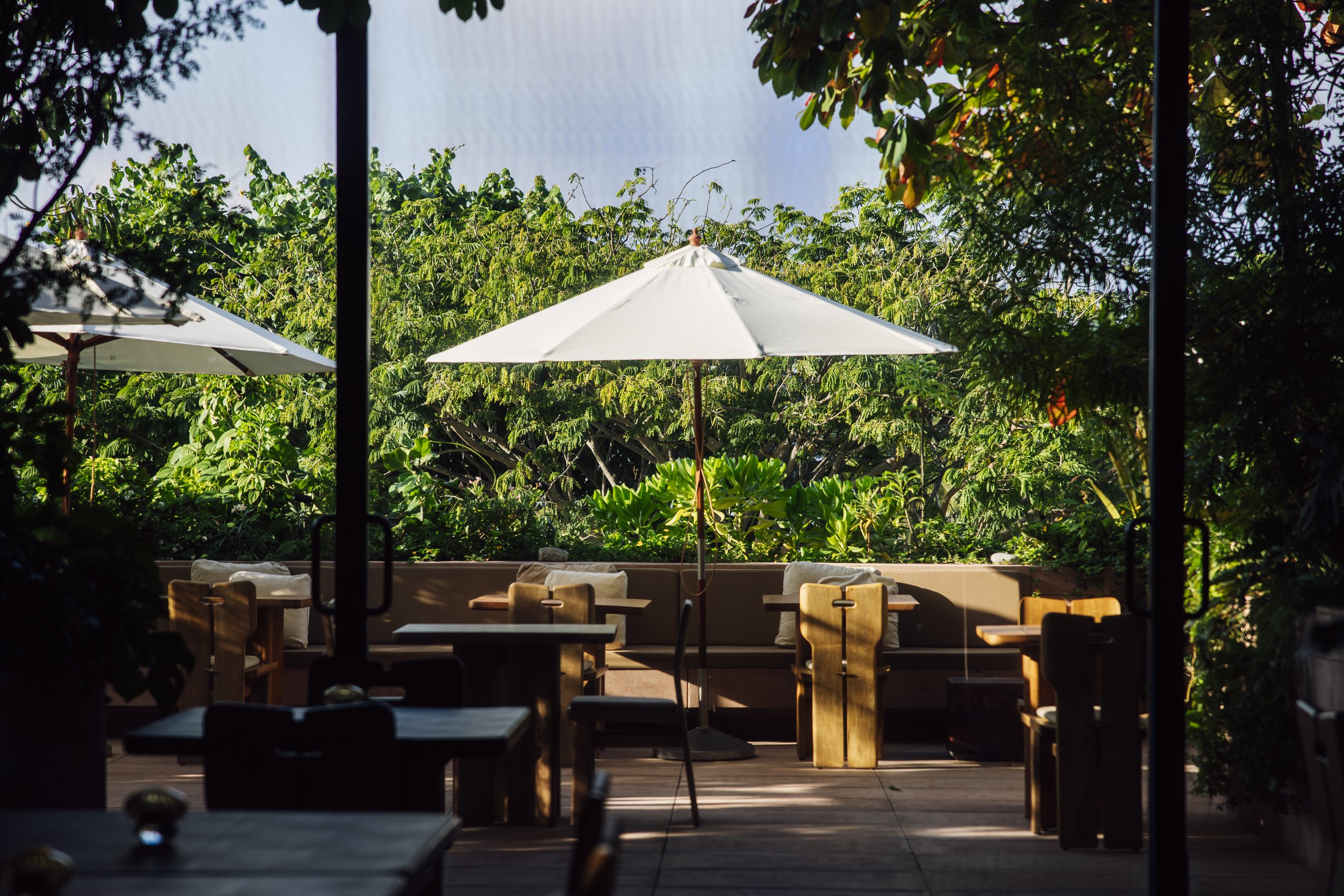 Outdoor patio with wooden tables and chairs, large white umbrellas, lush green trees in the background, viewed from inside through a doorway.